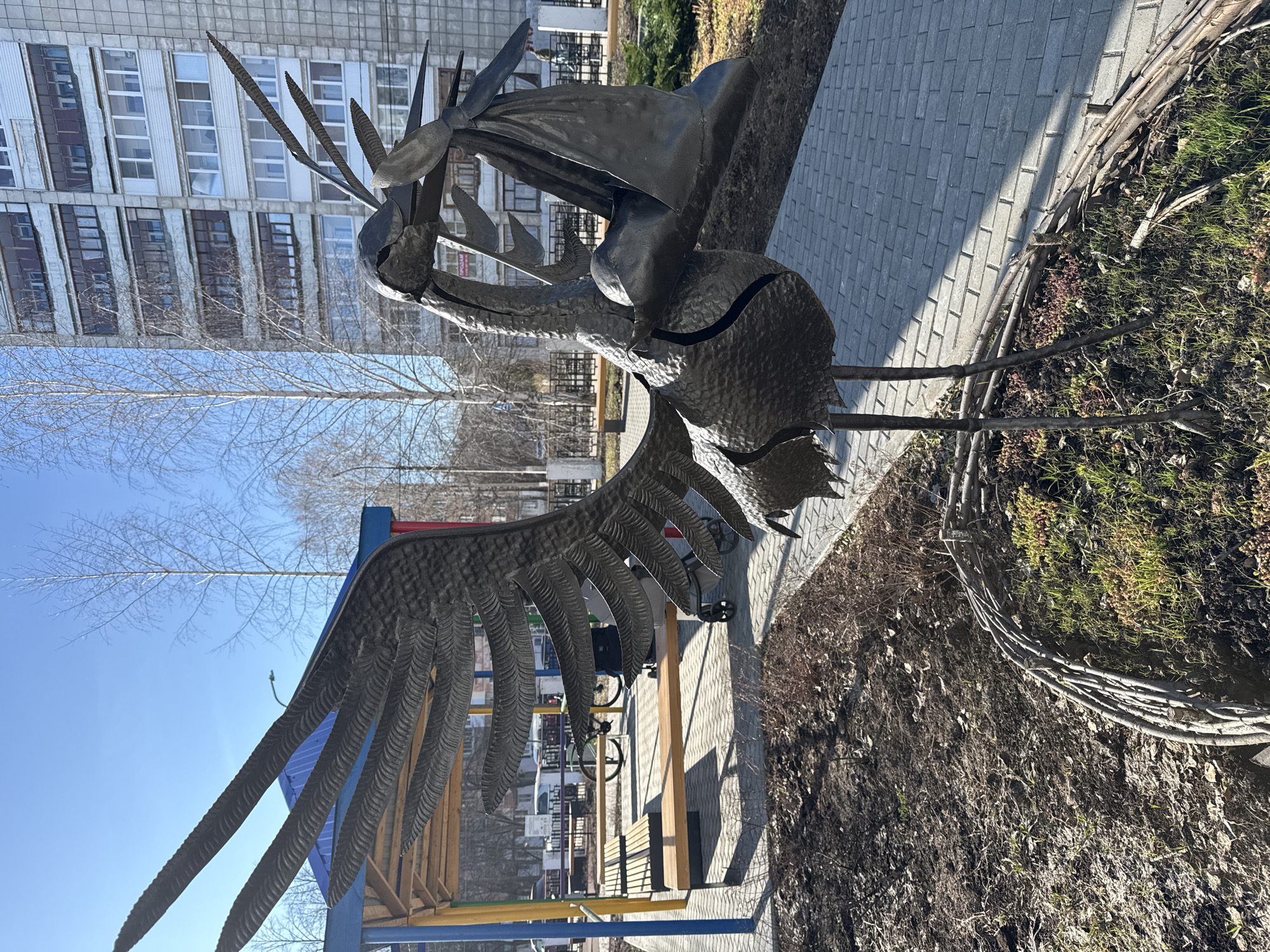 A metal sculpture of a bird-like figure, possibly a stork carrying something, stands in an outdoor setting in Unknown, Unknown. The sculpture is placed near a brick walkway and some greenery. In the background, a tall building with multiple windows can be seen under a clear blue sky. A playground structure is visible to the lower-left of the sculpture.