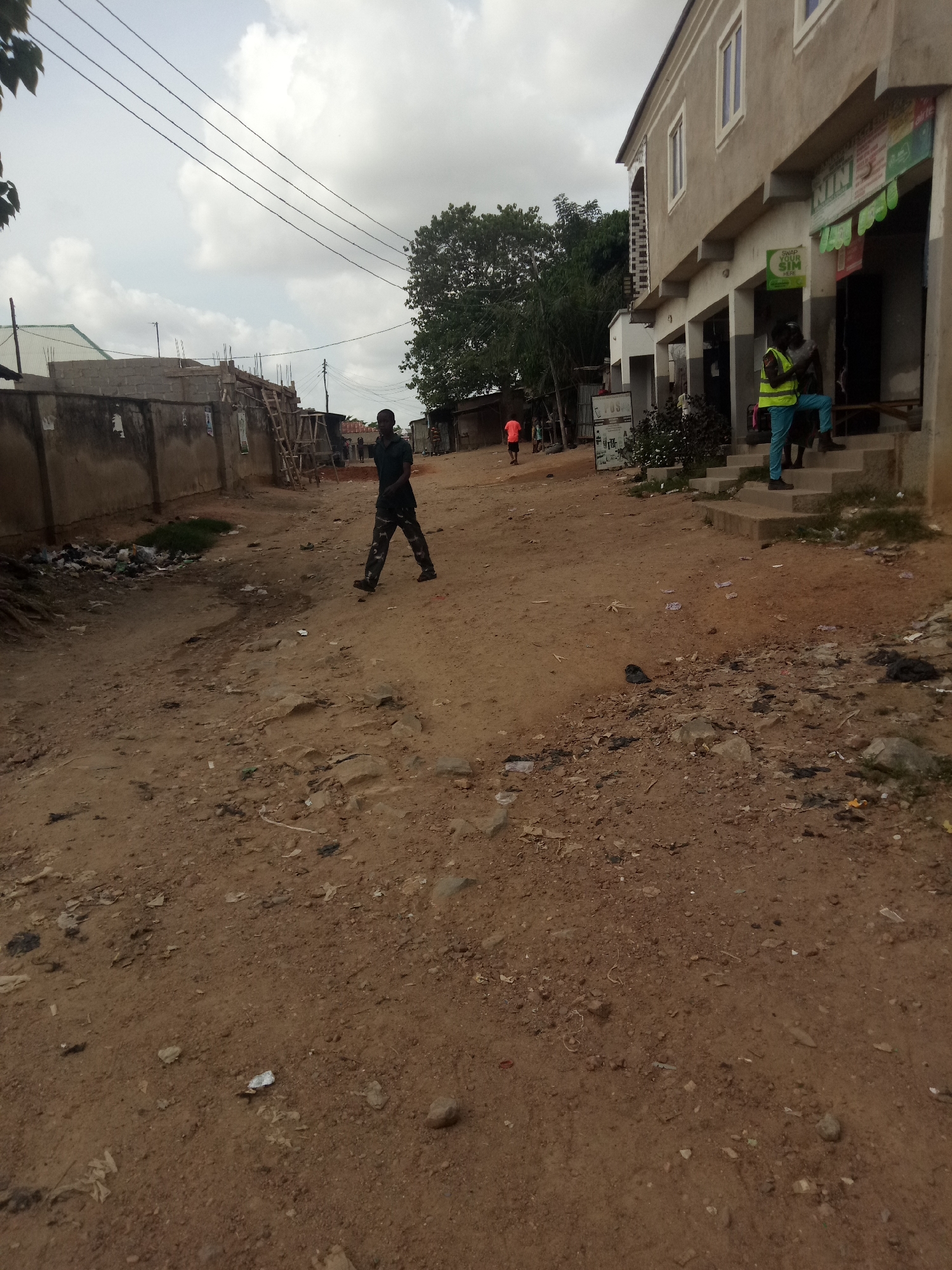 A male pedestrian walks on a dirt road in Uke, Nigeria, under a cloudy sky. The man, wearing camouflage pants and a dark short-sleeved shirt, is in motion, mid-stride. To his right, a two-story building with shops on the ground floor stands beside the road. Two individuals, one wearing a yellow safety vest, are visible near the building's entrance. The environment appears somewhat unkempt, with visible litter and debris scattered along the dirt road and near a low wall on the left side of the image. The road is unpaved and dusty. Overhead, power lines are strung across the sky. A small pile of garbage is visible against the wall on the left.