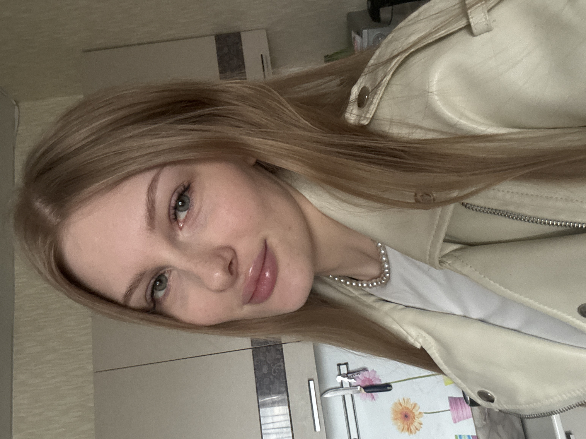 A young woman is featured in a close-up portrait, captured indoors. She has long, straight blonde or light brown hair and fair skin. Her green or hazel eyes are directed towards the camera, and she wears a soft, pleasant expression with a slight smile and glossy lips.
She is dressed in a light-colored, possibly cream or off-white, leather jacket featuring visible snap buttons and zippers. A white top is visible underneath her jacket, and she accessorizes with a pearl necklace.
The background suggests an indoor domestic setting, specifically a kitchen located in Кондратово, Russia. Visible elements include light-colored kitchen cabinets or cupboards with horizontal handles, and a part of a white refrigerator. The refrigerator is decorated with floral magnets, showing orange and pink flowers, and has a small knife magnetically attached to its surface. Other indistinct household items are faintly visible in the background.
The woman appears to be posing for the camera, likely taking a selfie, conveying a calm and friendly demeanor. No specific time of day or weather conditions can be determined from the indoor lighting. There is no visible text within the image.