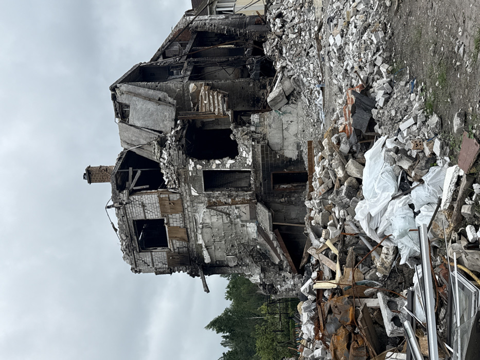 A severely damaged building stands against a cloudy sky. The structure is partially collapsed, exposing inner rooms and charred sections. Piles of rubble and debris surround the base, including fragmented concrete, twisted metal, and white plastic bags. Trees are visible behind the wreckage. The scene suggests a place affected by war or a major disaster.