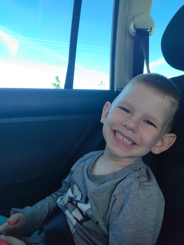 A young boy with short, light hair and prominent ears is captured in a close-up, smiling broadly at the camera. He is wearing a gray long-sleeved t-shirt with a dark graphic design visible on the front. His wide smile reveals his upper teeth, and a small gap or missing tooth is noticeable among his front teeth, indicative of a young child. His eyes are bright and joyful, matching his happy expression.
The setting is the interior of a vehicle, likely a car, seen from the back seat. A dark-colored seatbelt crosses his lap and chest, suggesting he is secured for travel. To the boy's left, a large car window reveals a bright, clear blue sky with a few faint white contrails or very thin clouds. A distant, blurred glimpse of green foliage, possibly a tree or bush, can be seen outside the window near the bottom. The car's interior appears dark, contrasting with the bright outdoor scene. A part of the car's headrest is visible in the upper right corner, and the dark upholstery of the car seat is also prominent.
The image conveys a sense of happiness and contentment, possibly during a daytime car journey on a pleasant day. No visible text is present in the image.