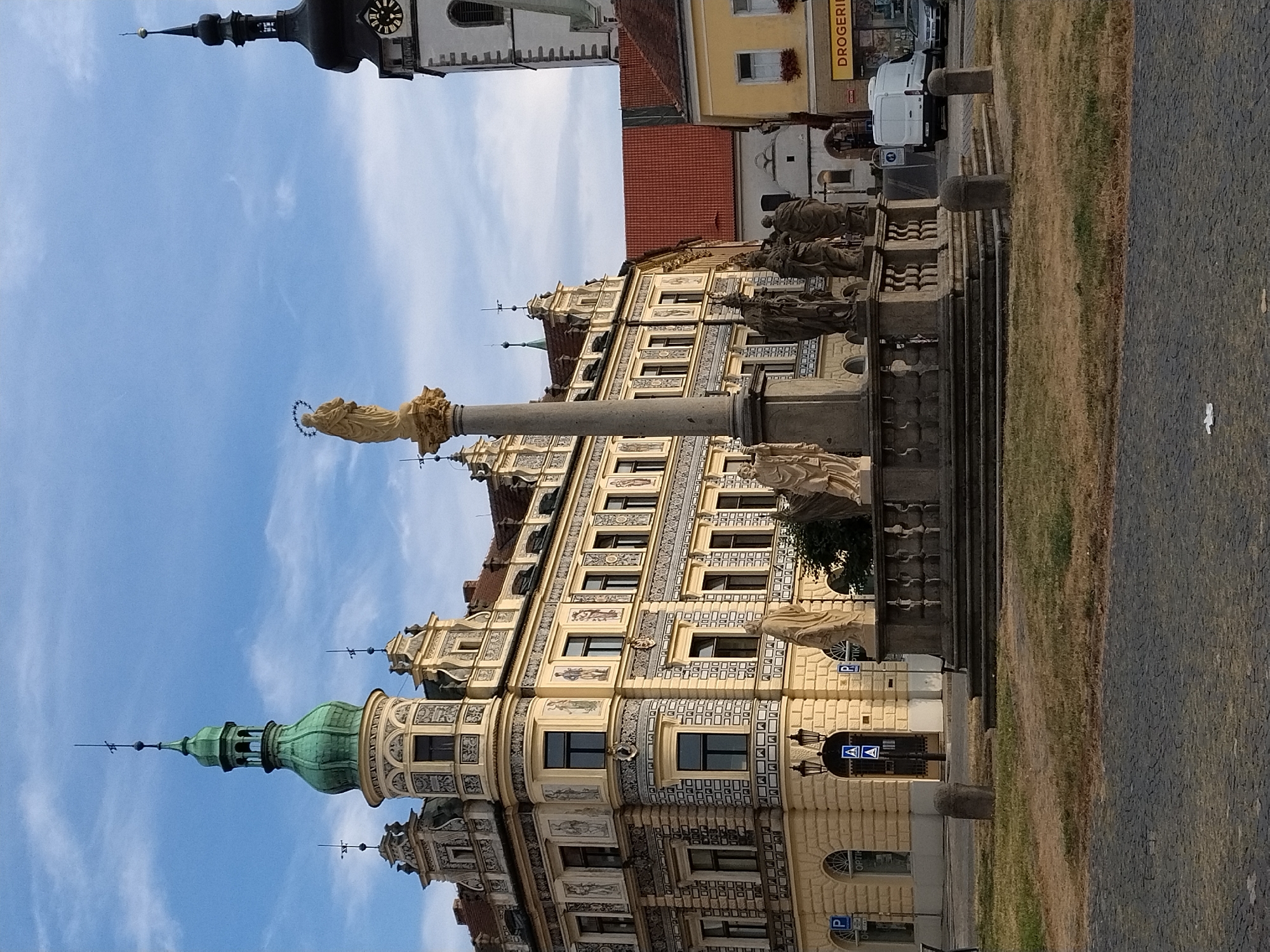 The image shows the central square of Písek, Czechia, featuring a Marian column adorned with statues and a statue of the Virgin Mary at its apex. The column is surrounded by a balustrade and steps. Behind the column, an ornate, cream-colored building with decorative elements and a green-domed tower is visible. To the right, a white van and a car are parked on the street in front of a yellow building with the word "DROGERIE" on its facade. A tall, dark church tower with a clock face is also present in the background. The ground in the foreground is paved with cobblestones and a patch of dry grass. The sky is blue with some scattered clouds. There are no visible people in the image.