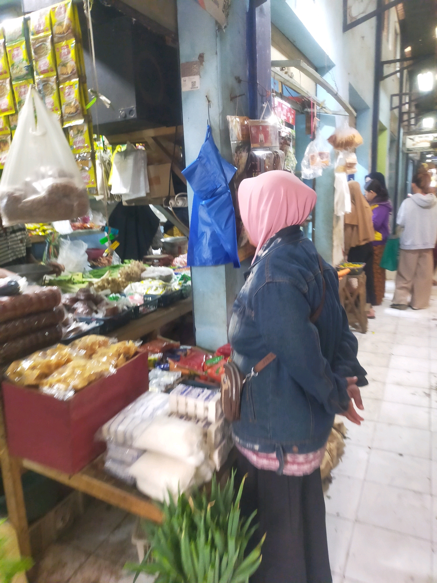 The image depicts a bustling market scene in Bululawang, Indonesia. A woman in a pink headscarf and denim jacket is standing, facing away from the camera, at a market stall. She is wearing a long black skirt and a brown crossbody bag. In the foreground, various food items are displayed on wooden tables. These include what appear to be crackers in a red box, bags of rice or sugar, and fresh produce like green onions. Behind the woman, other shoppers and vendors are visible, with market stalls displaying a variety of goods, including packaged snacks and fabrics. The market environment appears to be covered, with a blue plastic tarp hanging in the background and overhead lighting illuminating the area. The floor is tiled.