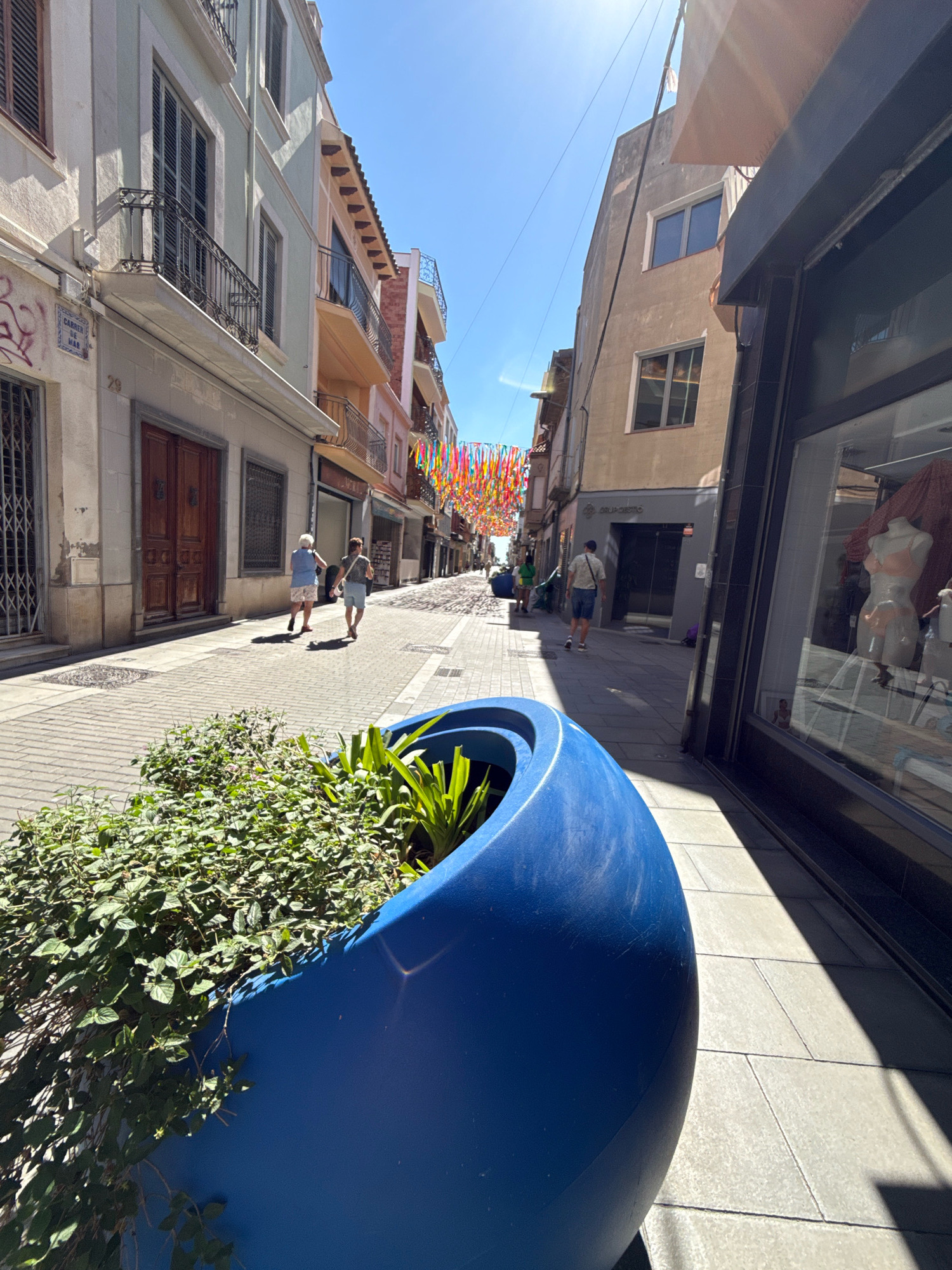 This image captures a sunlit street in Malgrat de Mar, Spain, likely during daytime. The perspective is low, looking down a pedestrianized street lined with multi-story buildings on both sides. In the foreground, a large, vibrant blue planter overflows with lush green foliage, partially obscuring the view of the sidewalk. The street itself is paved with light-colored tiles, with a distinct pattern of intersecting lines.
On the left side of the street, buildings feature traditional architecture with balconies and shutters. A street sign reading "CARRER DE MAR" is visible. Further down the street, two people are walking away from the camera, one in a blue top and patterned shorts, the other in a grey top and denim shorts.
The right side of the street is dominated by a dark-fronted building with a large shop window displaying mannequins dressed in swimwear. The window reflects the street scene. Further down this side of the street, a building with a beige facade has two windows.
Above the street, a canopy of colorful streamers stretches between the buildings, suggesting a festive atmosphere. People are seen walking further down the street in the distance, and the clear blue sky suggests good weather. The overall impression is of a lively, charming Mediterranean street.