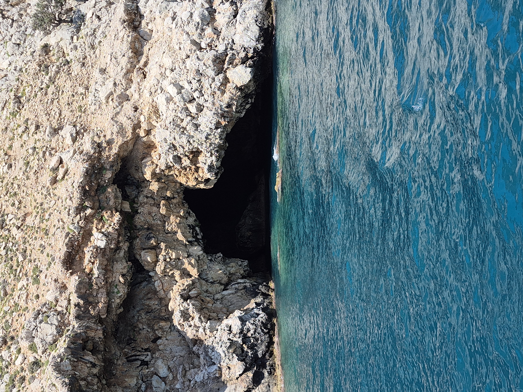 A vertical shot depicts a rocky cliff meeting the clear blue sea, possibly in Alanya, Turkey. The cliff face is composed of light-colored rock formations with patches of vegetation, while the water is a vibrant turquoise color marked with ripples. A dark cave is visible within the rock face, adding a sense of depth.