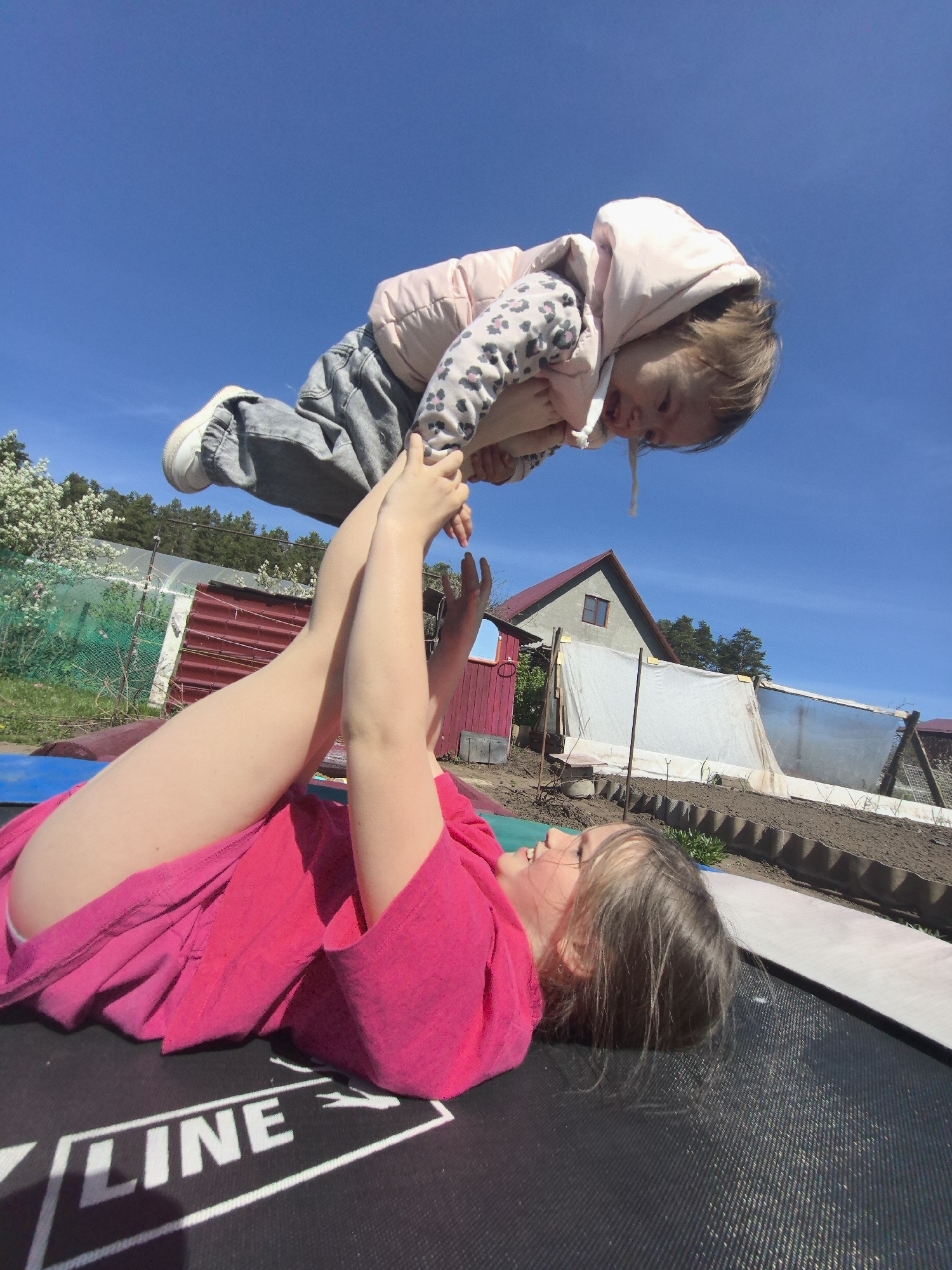 A woman is lying on her back on a trampoline, holding a young child in the air with her arms. The child is wearing a jacket with a hood and grey pants with white shoes. Behind them, there is a house and a garden area. The sky is blue. "LINE" is written on the black trampoline in white letters. Based on the landscape and architectural details, the scene may be located in a rural or suburban area. The location is Noviy, Russia.