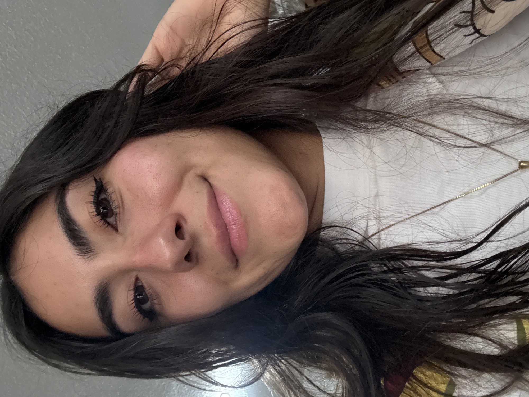 A female with dark hair is lying down, facing the camera. She has dark eyebrows, dark eyeliner, and appears to be wearing lipstick. She is wearing a white shirt and a necklace with a small pendant. The background is blurred. Location: Puebla, Mexico is not determinable from the image.