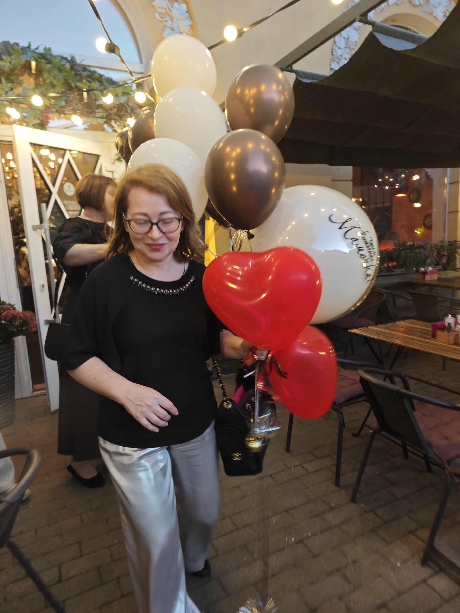 A woman wearing glasses and a black blouse with shiny silver pants stands outside a cafe, holding a bouquet of red and metallic balloons. She is smiling and looking down at the balloons. In the background, another woman in a black dress is visible. The scene appears to be a celebration, possibly a birthday or anniversary, taking place in the city of Pushkin, Russia, as indicated by the inscription on one of the balloons. The environment suggests an outdoor seating area of a restaurant or cafe, with tables and chairs visible. The lighting indicates it is likely evening, with string lights adding to the festive atmosphere.