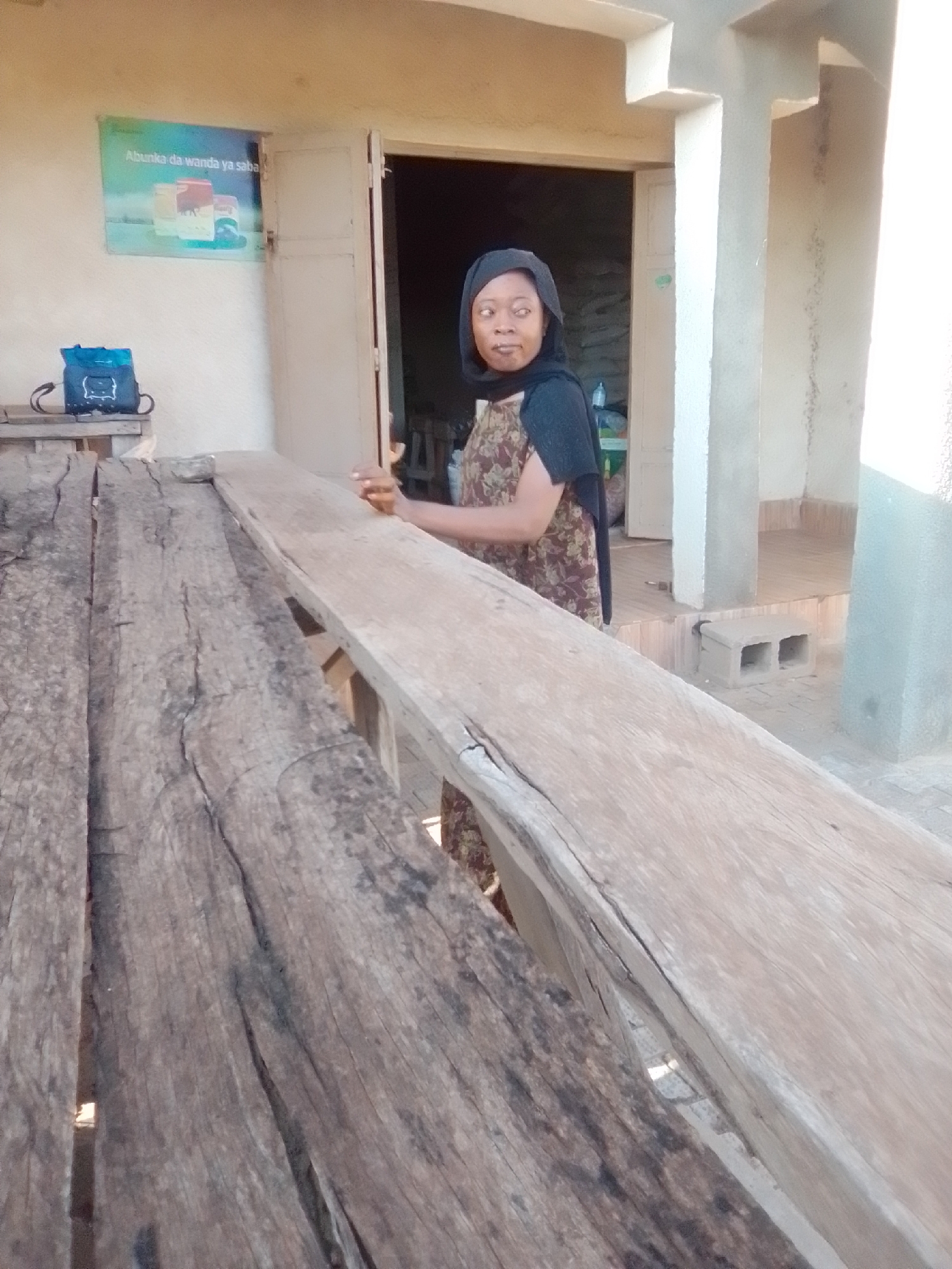 A woman wearing a black hijab and a patterned dress stands in the doorway of a building. She is looking to her right, with her mouth slightly open. To the left of the woman, a blue backpack rests on a wooden surface. The building appears to be made of concrete and has a light-colored facade. In front of the building is a long, rustic wooden table made of rough-hewn planks. The scene suggests a street-level view in a Nigerian city, possibly Galadima, given the context. The lighting indicates it is daytime, with shadows suggesting a strong sun.