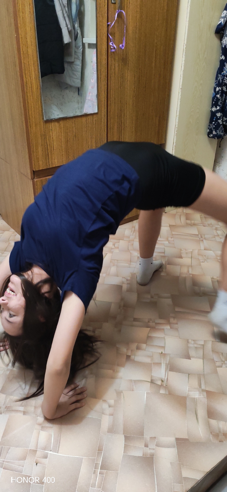 A woman performs a backbend on a tiled floor indoors. She wears a navy blue t-shirt and black shorts. Her dark hair hangs towards the floor. She is smiling. A wooden wardrobe stands in the background. The image contains "HONOR 400" at the bottom left corner. The location of the photograph is unknown.