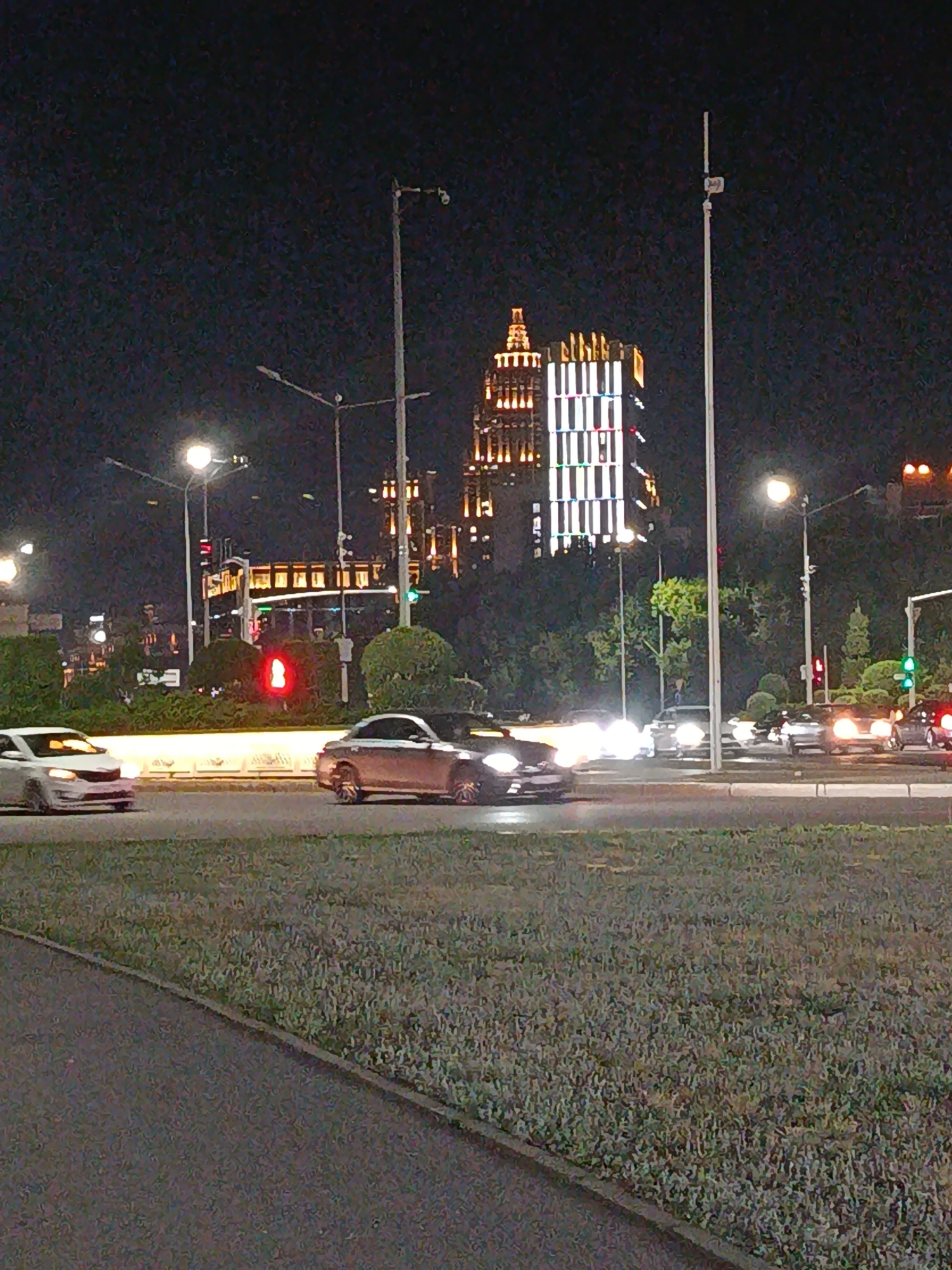 The image depicts a nighttime street scene in a city, likely an urban intersection with several lanes of traffic. Tall buildings with illuminated windows and decorative lighting are visible in the background, suggesting a developed commercial or residential area. Streetlights illuminate the road, and traffic signals are present, one of which is showing a red light. Several cars are on the road, some in motion and others stopped. One car in the foreground is white, while another is darker in color. Green landscaping with bushes and trees is present along the median or sidewalk. The overall impression is that of a busy city at night, with vehicles and buildings characteristic of a modern urban environment. The image quality is somewhat grainy due to low light conditions.