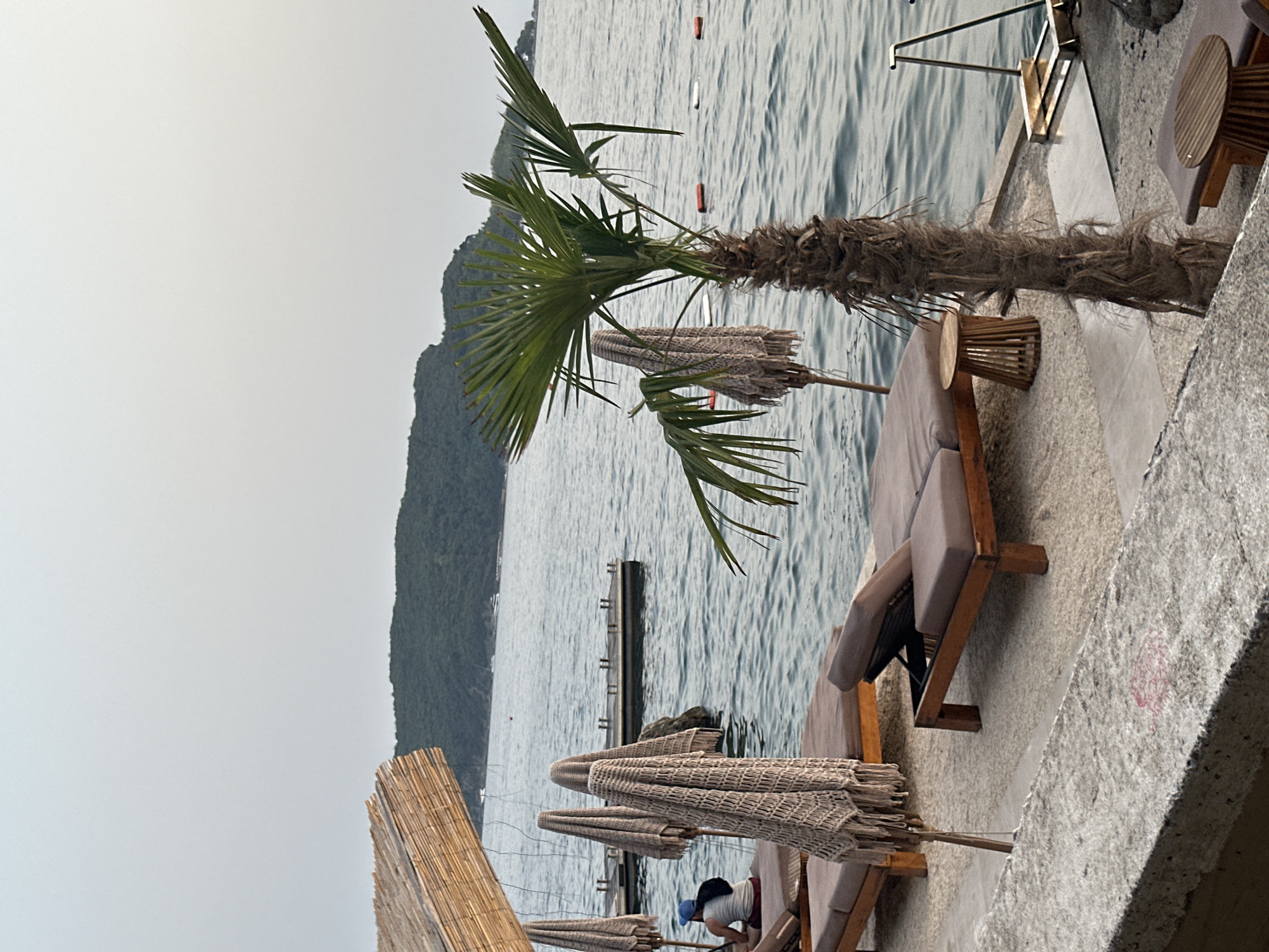 The image depicts a tranquil beach scene in Budva, Montenegro. In the foreground, there are several empty lounge chairs and a small wooden table, suggesting a relaxing atmosphere. A thatched roof overhang provides shade. In the middle ground, the calm sea stretches out, with a small pier visible in the distance. A lone palm tree stands prominently, adding to the tropical ambiance. The background features a lush green hillside, characteristic of the Montenegrin coast. The sky is overcast, with soft, diffused light. A person can be seen seated on one of the lounge chairs in the far left of the image. The overall scene conveys a sense of peace and serenity.