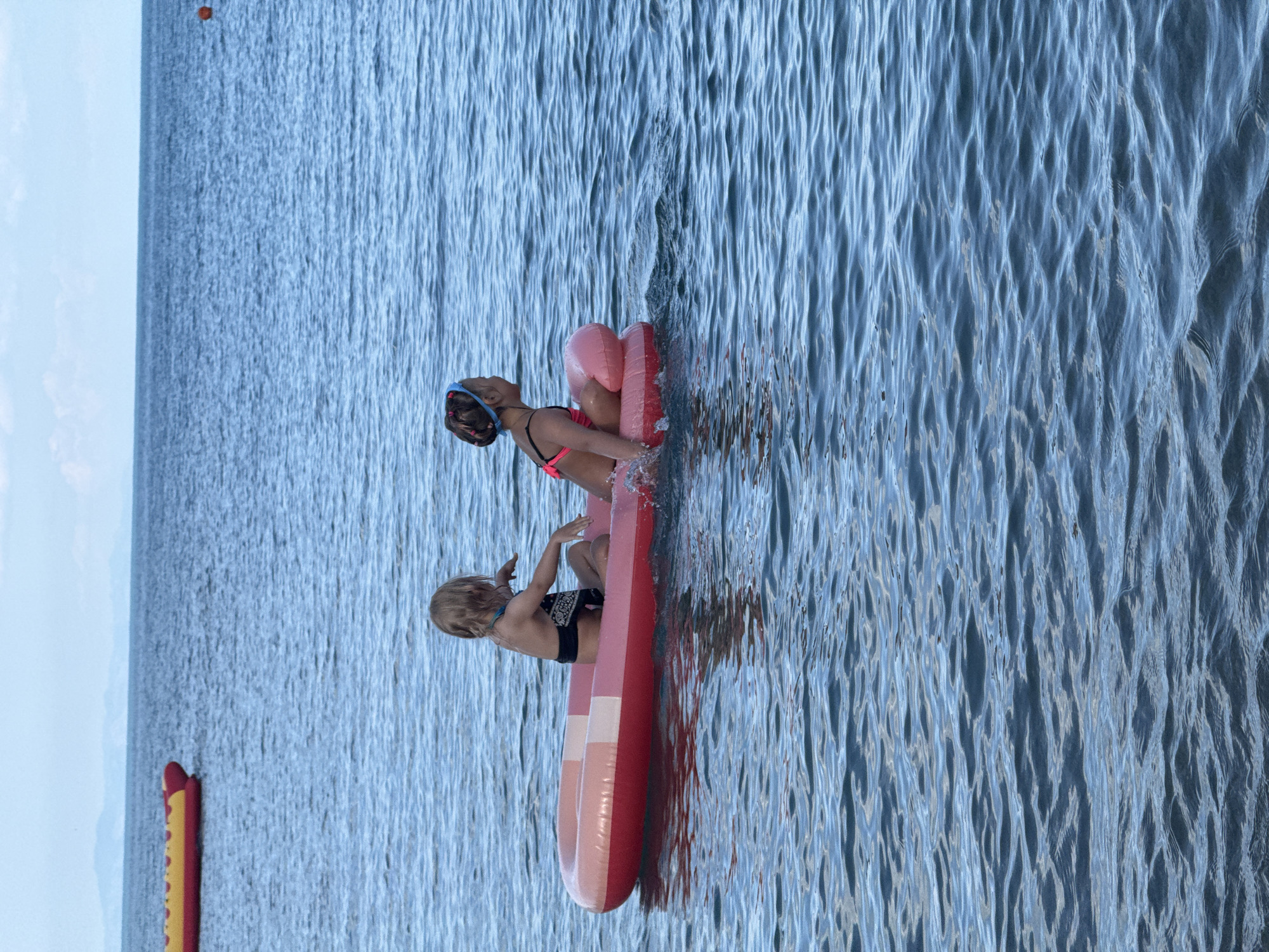 Two young children are playing on a pink and red inflatable in the water. The child on the left is wearing a blue swimming cap and a pink bikini top, and appears to be splashing. The child on the right is wearing black swim trunks and has blond hair. They are both sitting on the inflatable. The water is calm with some ripples. In the distance, a yellow and red inflatable object is visible. The sky is overcast. The scene appears to be a recreational area, possibly a lake or the ocean. The children seem to be enjoying themselves. There is no visible text in the image.