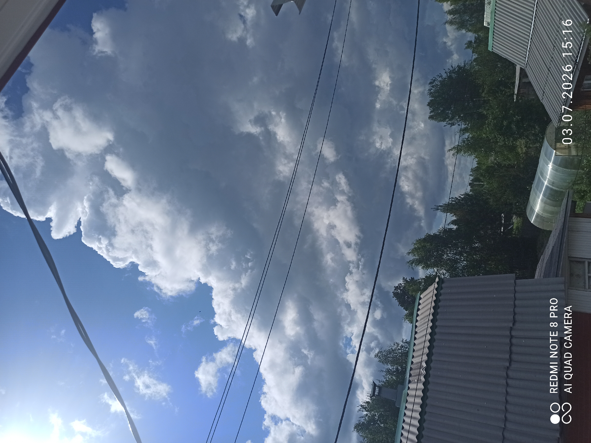 The image depicts an outdoor scene with a vast, cloudy sky. The clouds are a mix of dark and light gray, with patches of bright blue sky visible. The sun is shining brightly from the upper left corner, casting a lens flare across the sky. Several power lines are stretched across the frame, with one prominent line crossing diagonally from the top left to the bottom right. Below the sky, a line of trees is visible, with dense green foliage. In the foreground, the corrugated gray roof of a building is prominent, with a section of a greenhouse visible to the right. The image includes a timestamp in the lower right corner: "03.07.2026 15:16" and a watermark in the lower left corner indicating "REDMI NOTE 8 PRO AI QUAD CAMERA". The scene appears to be a residential area, possibly in a rural or suburban setting, given the presence of trees and simple structures. The city of Sovetskiy, Russia is not directly identifiable from the visual elements in the image.