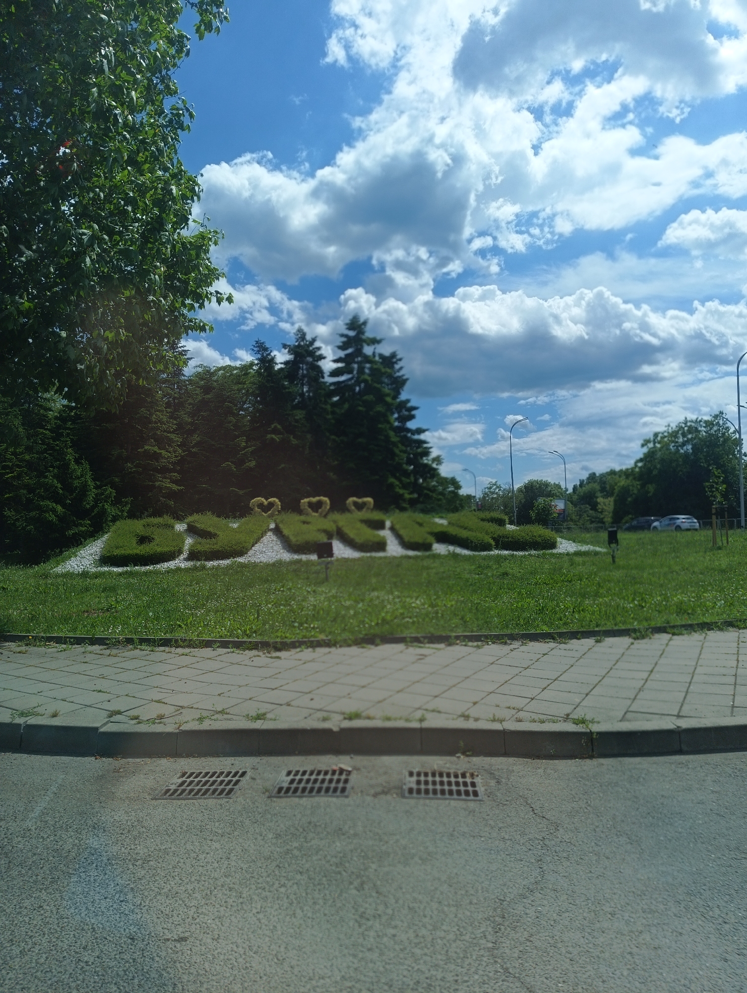 The image shows a grassy area with topiary spelling out "BURGAS" with heart shapes above some letters. The topiary is set against a backdrop of trees and a bright blue sky with white clouds. In the background, there are streetlights and some parked cars. The foreground features a paved sidewalk and a street with drainage grates. This scene is likely located in Burgas, Bulgaria, given the name spelled out in the topiary. There are no people visible in the image.