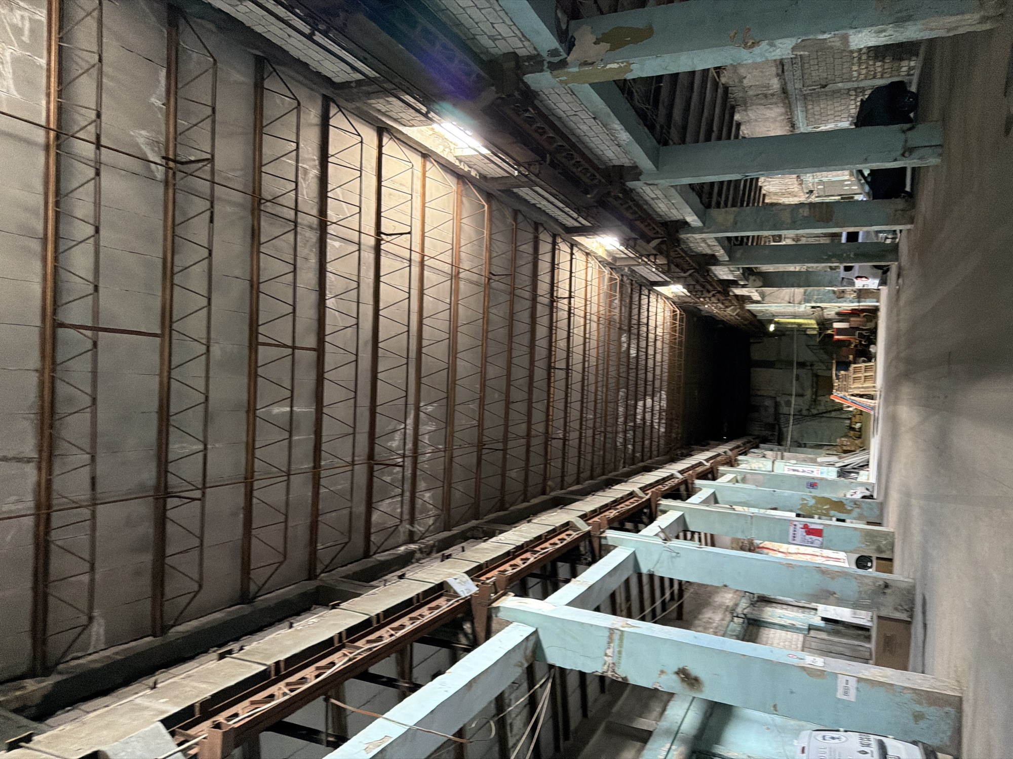 The image depicts a long, narrow, industrial-looking space. The walls are lined with a grid-like structure. A rusted metal track runs along the floor, partially covered by light gray slabs. Above, there are horizontal beams, painted in light blue. The end of the space recedes into darkness. People can be seen in the distance, obscured by the shadows.
Based on the image, it is not possible to confirm the location in the city of Yerevan, Armenia. There is no information in the image that would indicate its exact location.
