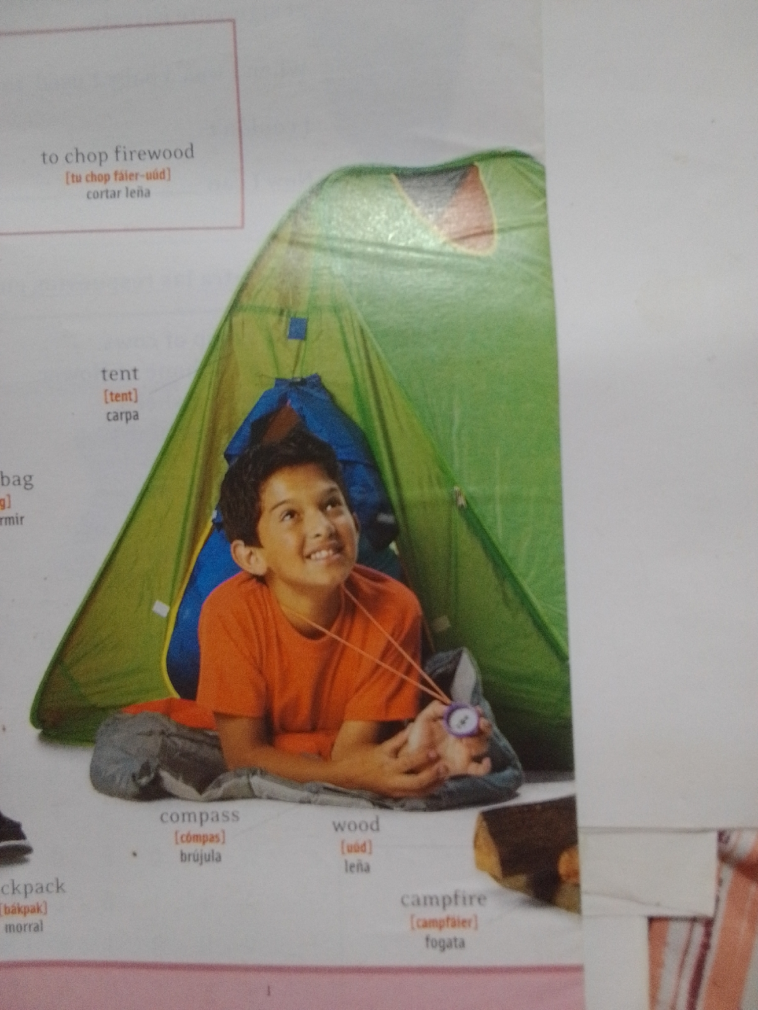 The image shows a young boy inside a green tent. He is smiling and looking up, holding a compass in his hand. Next to him is a blue sleeping bag and a backpack. In the foreground, there are labels in English and Spanish for various camping-related items, including "tent," "compass," "wood," "backpack," and "campfire." There is also a log, presumably for a campfire, visible in the lower right corner. The image appears to be from an educational book or resource. There is no information in the image or the provided crops that places the scene in La Tagua, Venezuela.