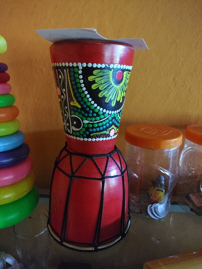 The image presents a still life featuring a prominent, decorative drum-like object in the foreground, set against a warm, orange-yellow wall. The central object is a red, hourglass-shaped drum, possibly a djembe. Its upper, tapering half is painted black with vibrant, intricate patterns in green, yellow, pink, and white dots and lines, resembling folk or aboriginal art. A piece of light-colored cardboard or paper rests on the top opening of the drum. The lower, wider section of the drum is also red, and features dark lacing or cord tied around it in a geometric pattern, connecting to a light-colored rim at the base.
To the left, a colorful stacking toy is partially visible, displaying rings in shades of green, yellow, pink, blue, and orange. To the right, two clear plastic containers with bright orange screw-on lids are present. The container closer to the drum appears to hold small objects, including a yellow toy and some white, spoon-shaped items. A stack of small, rectangular cards is also visible behind the containers.
The items are arranged on a dark, reflective surface, likely a table or shelf. The overall setting appears to be indoors, possibly a domestic environment or a small shop. There is no visible text or specific activity taking place; it is a static display of objects. The image is well-lit, suggesting daytime indoor conditions.