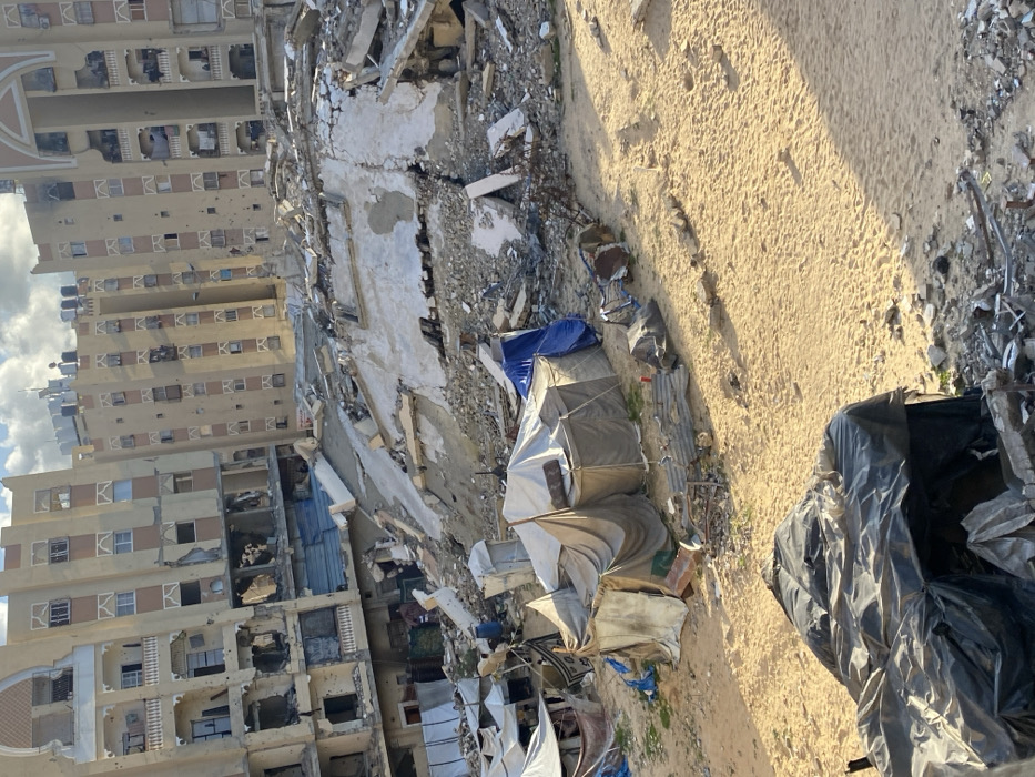 Extensive damage is evident in a densely populated area. A high-rise building shows signs of heavy impact with multiple damaged windows and structural compromise. Rubble and debris are scattered across the ground, creating an uneven landscape. Makeshift tents and shelters stand among the ruins, suggesting displacement and emergency housing. Black plastic sheeting is visible, potentially used for protection or waste containment. The scene evokes a sense of destruction and humanitarian crisis.