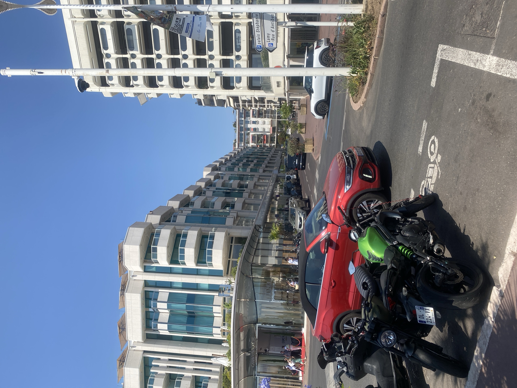 In Cannes, France, on a sunny day, a street scene unfolds with modern architecture and parked vehicles. Several multi-story buildings with white facades and numerous balconies line the street. The buildings feature a mix of glass-fronted sections and rows of rounded windows. A prominent white building on the right has a modern, tiered design.
In the foreground, two motorcycles are parked on the right side of the street. One is dark-colored, and the other is bright green. To their right, a red convertible car is parked, facing away from the viewer. Further down the street, a white compact car is visible.
On the left side of the image, a building with a glass facade appears to be a commercial establishment, with people visible inside and on a red carpet leading to the entrance.
Street signs are present, including one with directional arrows indicating "palais Stéphanie" and "Lamy - Rue d'Antibes". A lamppost with a security camera is also visible. The sky is clear blue.