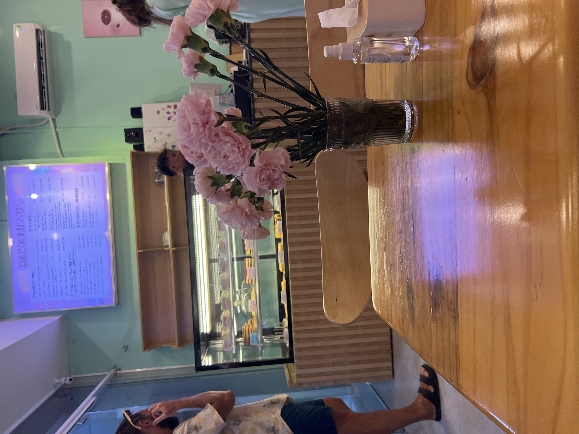 Here's a concise description of the image:
The image appears to be taken inside a cafe or restaurant. The scene is dominated by a wooden table, on which sits a vase filled with pink carnations, a bottle of hand sanitizer, and a tissue dispenser.
In the background, there is a refrigerated display case filled with pastries or food items. There's also a menu board visible on the wall, displaying drink options in both English and likely Vietnamese. People can be seen: one standing behind the refrigerated display case and another partially visible at the bottom of the frame, appearing to be looking at a phone or something in their hand.
Based on the OCR text on the menu, there is a chance that this photo could have been taken in Bac Nha Trang Ward, Vietnam.