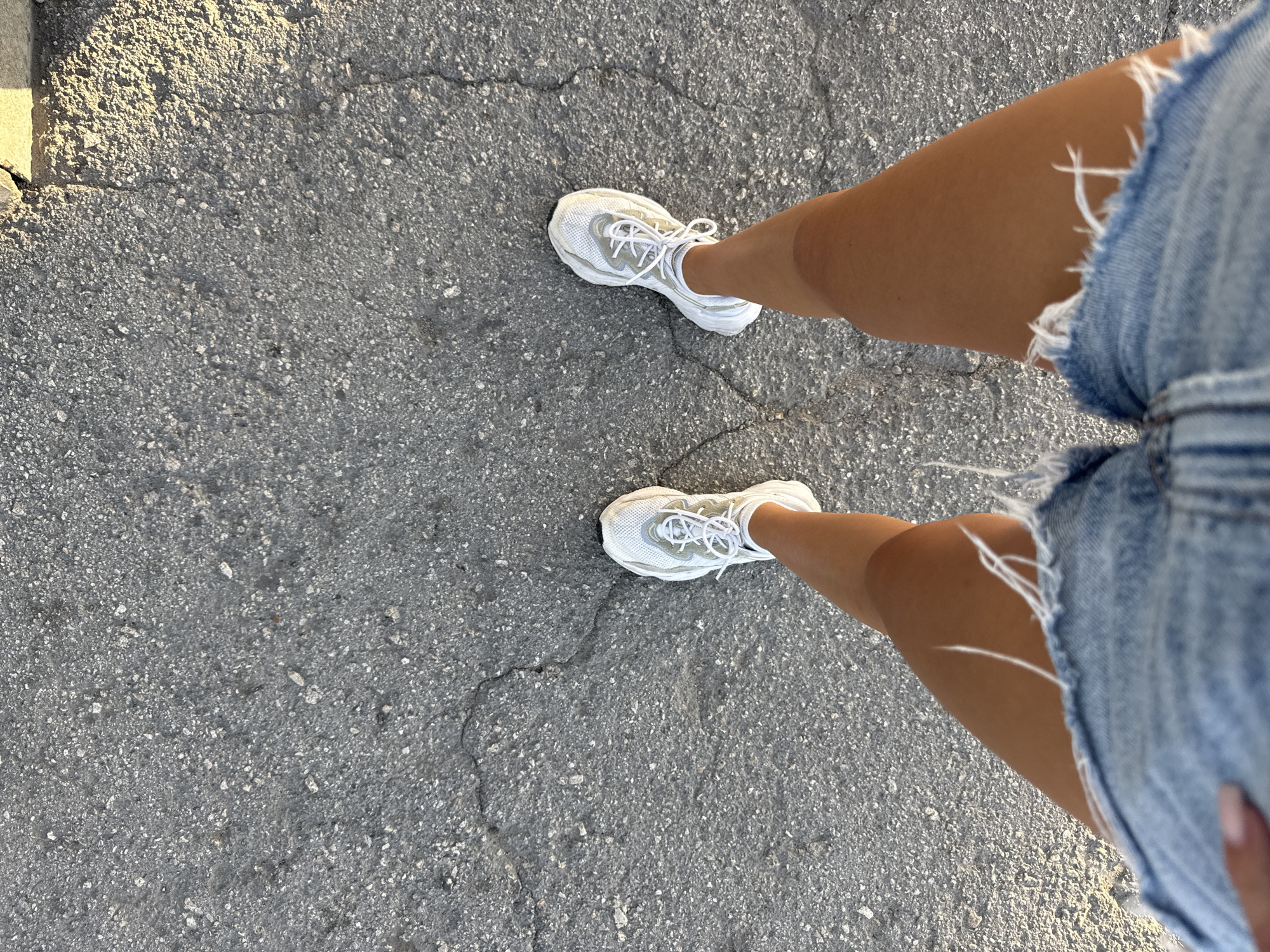 A top-down perspective shot shows a person standing on a cracked asphalt surface, likely a street or sidewalk. The person is wearing distressed denim shorts and white athletic shoes. The lighting suggests it is either early morning or late afternoon, with shadows cast by an unseen object to the right. The context of Cimișlia, Moldova, is noted, but no specific landmarks are visible to confirm this location. The scene is devoid of other people or significant urban features, focusing solely on the individual's attire and the ground beneath them.