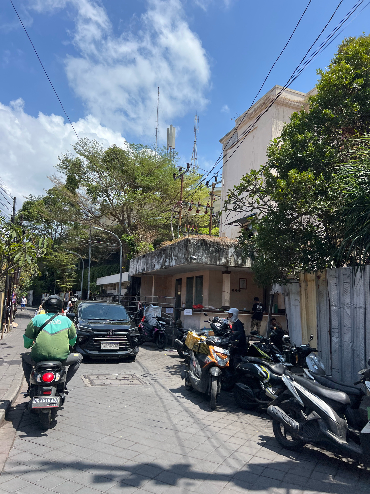 In Kuta, Indonesia, on a sunny day, a street scene unfolds with a mixture of vehicles and buildings. A dark-colored car is parked in the middle of the road, with its license plate visible. Several motorcycles are also present, some parked along the side and others in use. One person is riding a scooter, wearing a green and blue jacket and a black helmet. The street is paved with square cobblestones, and shadows from the buildings and trees fall across the ground. Buildings with light-colored facades line the street, with some featuring balconies and overgrown vegetation. Power lines crisscross the sky, and a cell tower is visible in the background. The environment appears to be a commercial or residential area with a moderate amount of activity.