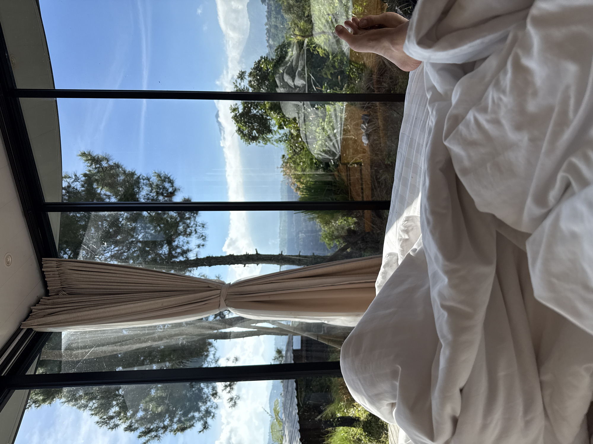 The image depicts a view from inside a building, likely a bedroom, in Kintamani, Indonesia. The foreground shows a white bedsheet bunched up on the right, with a bare foot visible. A large window with black frames dominates the view, showcasing the surrounding landscape. The window is partially covered by light brown curtains tied to the side. Outside, there are lush green trees and foliage, characteristic of the Kintamani region. The clear sky suggests a sunny day. The reflection in the window shows the outdoor scene, including a mountain range with a faint presence of a settlement in the distance.
