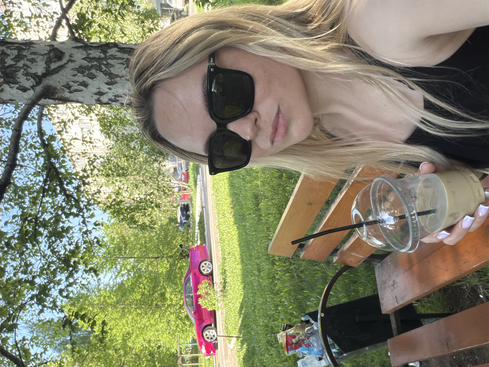 A woman with long, blonde hair and sunglasses is sitting at a wooden picnic table outdoors, holding a plastic cup with a straw in her right hand. The cup appears to contain a light-colored beverage, possibly iced coffee. She is wearing a black top. A tree with white and grey bark is visible to the left. In the background, there is green grass and a pink car parked on the side of a street. Based on the vegetation and typical urban features of Votkinsk, Russia, the location may be a park or recreational area within the city. The picnic table is located next to a metal trash can filled with plastic water bottles. The woman appears to be taking a selfie or has angled her phone to take the photograph.