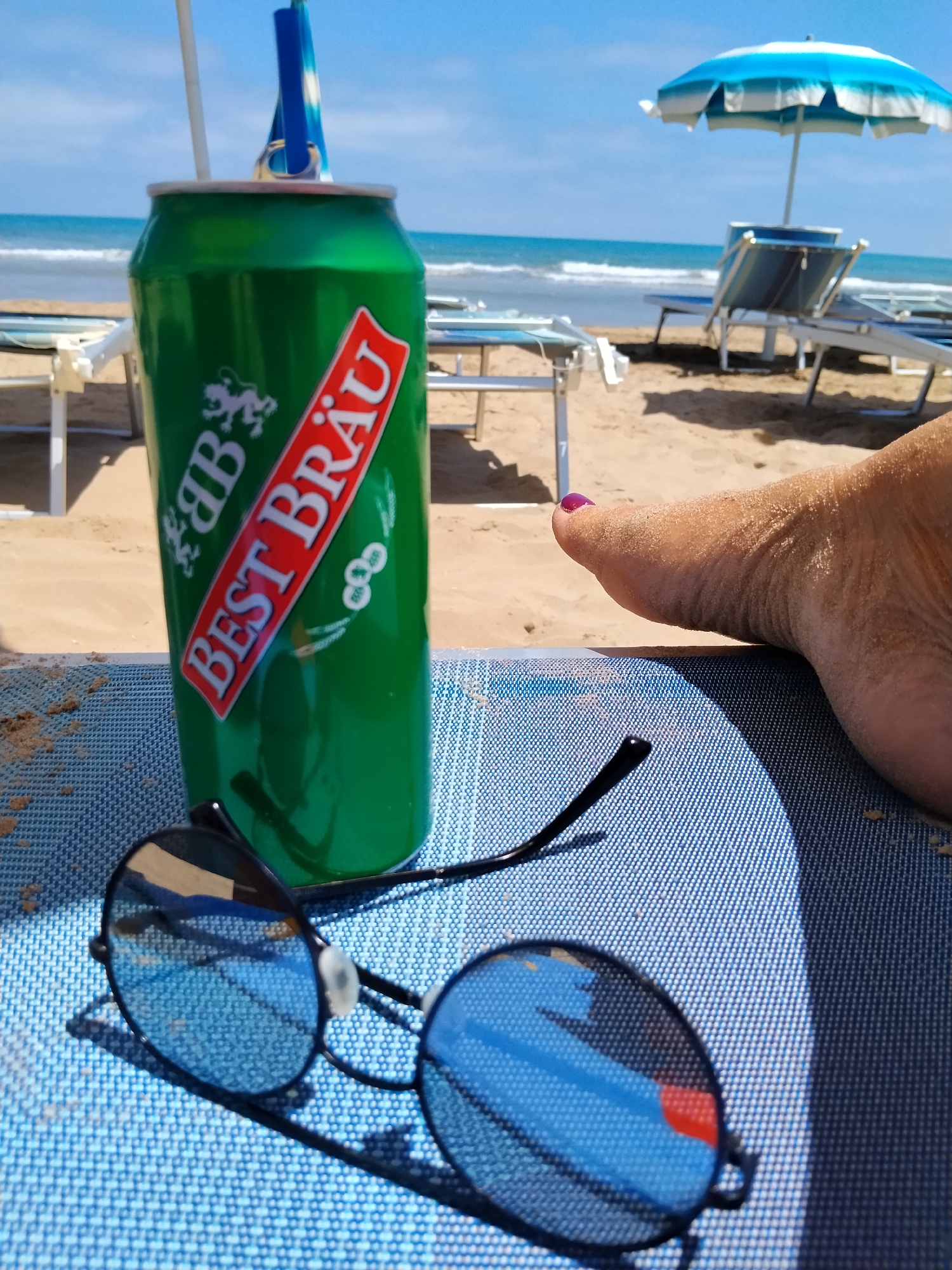 The image depicts a close-up view of a green beer can with the "BEST BRÄU" logo, a pair of sunglasses, and a sandy foot with purple nail polish, all placed on a blue, textured surface. In the background, a beach scene unfolds with lounge chairs, a blue and white umbrella, and the sea with gentle waves under a clear sky. The overall impression is a relaxed beach setting.