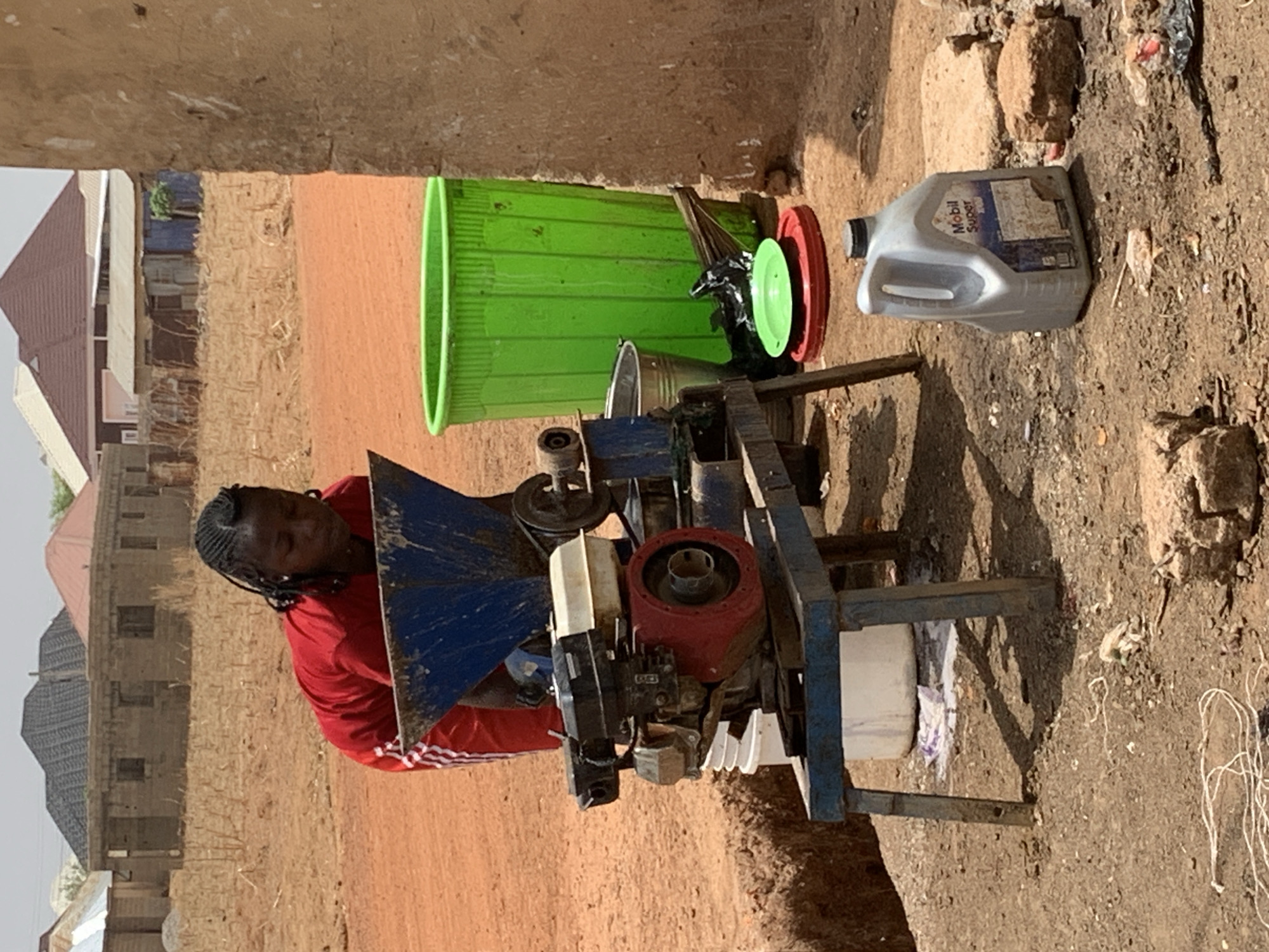 In Rafin Bauna, Nigeria, an African woman in a red shirt stands beside a mechanical device, possibly a grinder or mill. The device is constructed of blue metal, white plastic, and a red engine component. Near the device, there's a green plastic bucket and a Mobil Super oil container. The environment is a dirt ground with scattered debris, adjacent to buildings.