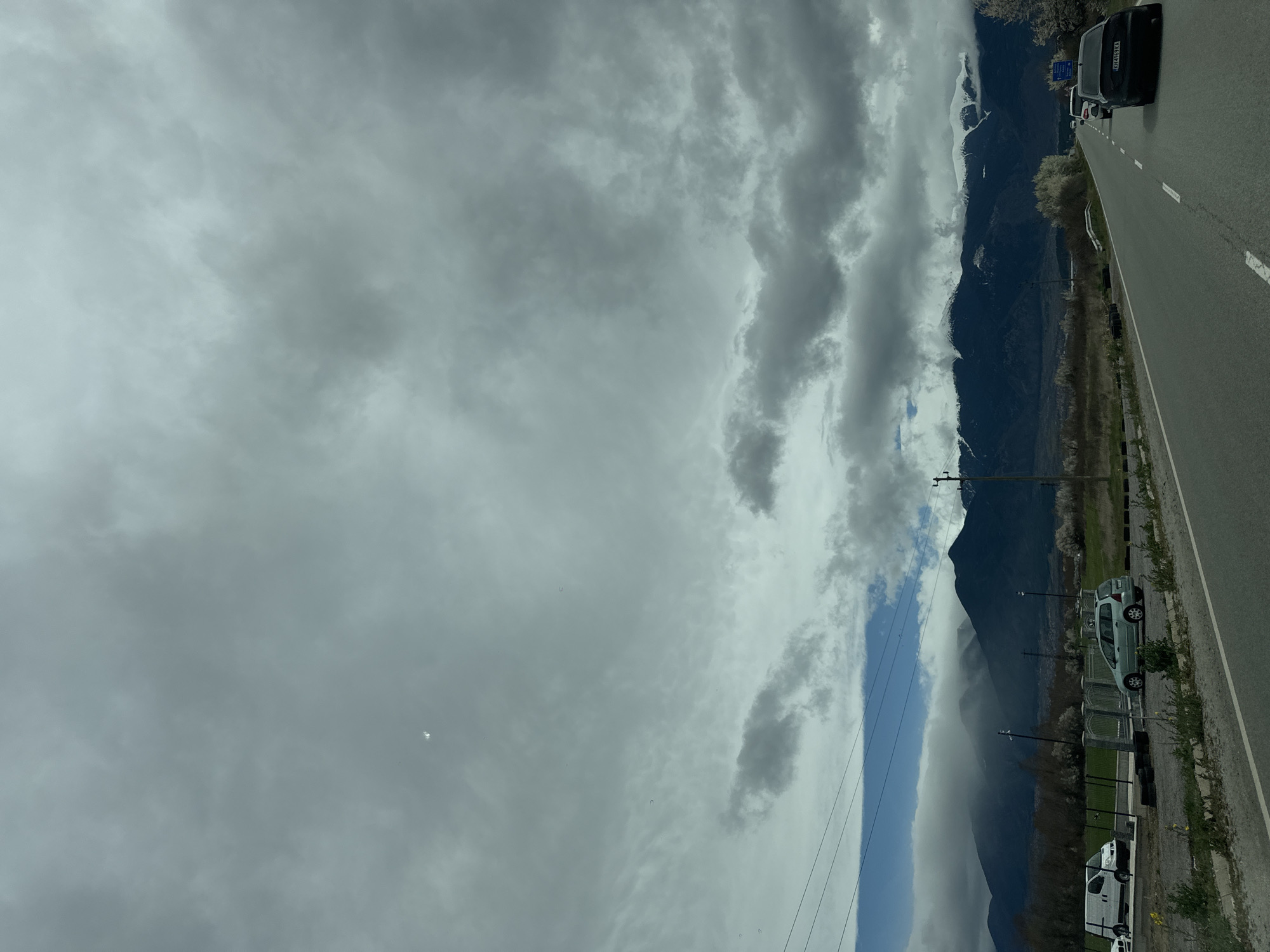 The image shows a road next to a mountain range under a cloudy sky. A black SUV is parked on the right side of the road. A small green car and a white van are also parked further down the road. Utility poles and trees are present on the mountainside. The scene is likely in Blatino, Bulgaria, due to the mountains. The sky is mostly overcast.
