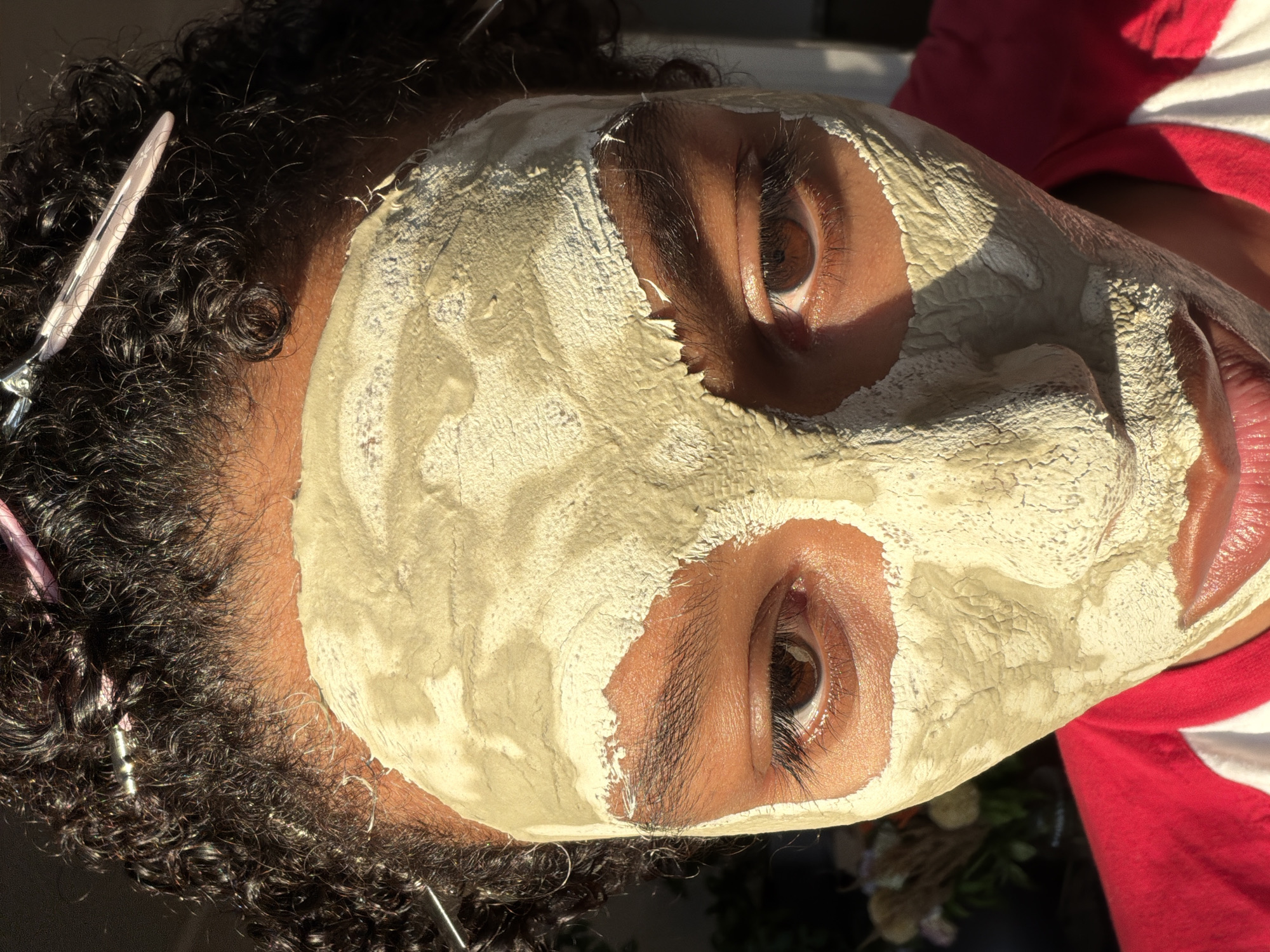 A close-up, head-on shot shows a person's face covered in a light beige, textured clay mask. The mask is applied unevenly, with some areas cracked and dry, leaving the eyes and mouth exposed. The person has dark, curly hair with several silver and pink hair clips holding it back from their forehead. The lighting suggests an outdoor setting, with harsh sunlight casting shadows on the face and highlighting the texture of the mask. The person is wearing a red and white t-shirt. No other details about the location in Champs-sur-Marne, France, are discernible from the image.