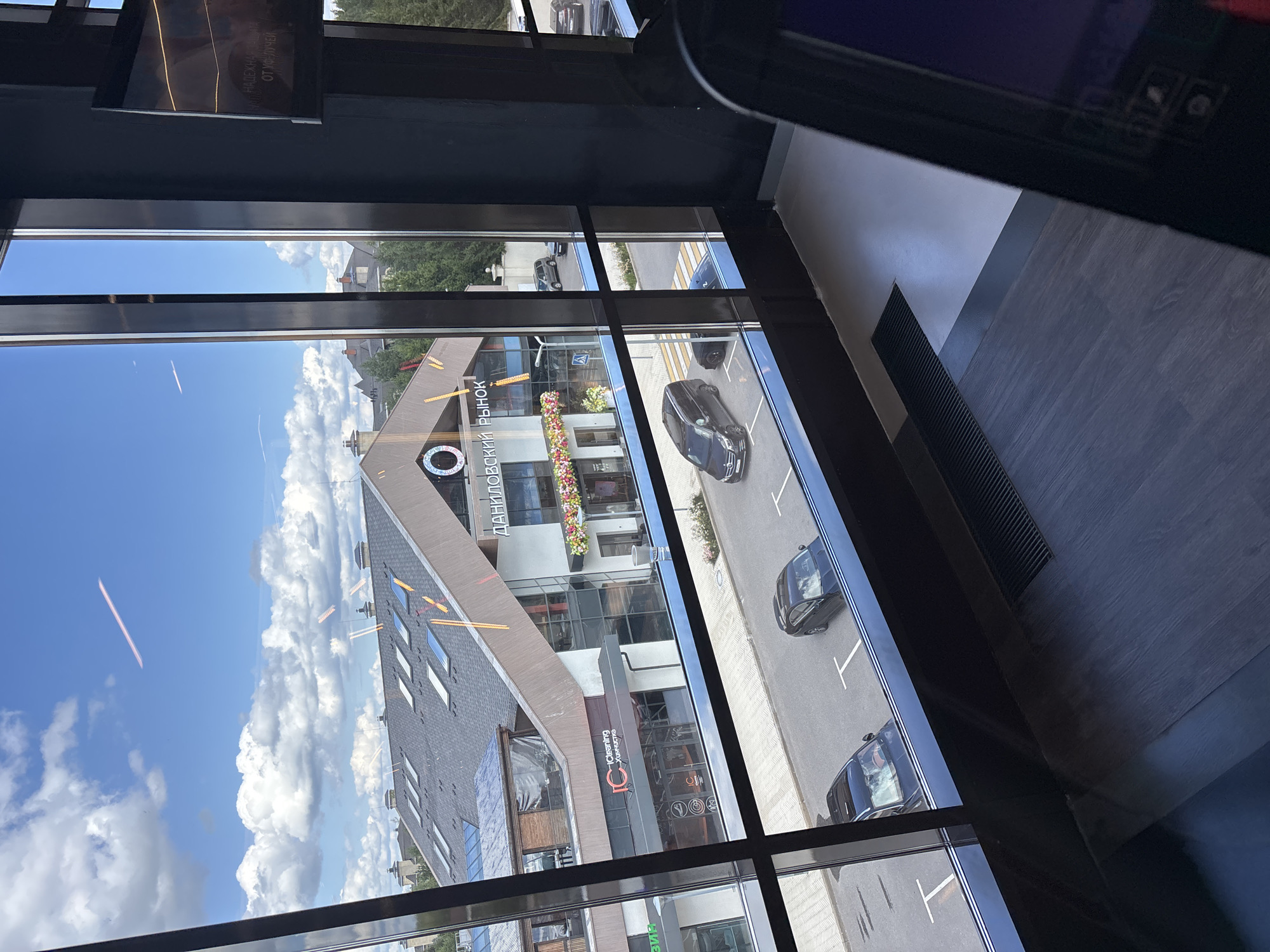 The image captures a view through a large window, likely from an interior space, looking out onto a street and buildings under a cloudy sky. The architecture suggests a modern, possibly commercial area.
In the foreground, the window frame and a dark, reflective surface of what appears to be a piece of furniture or equipment are visible. The window itself has dark, vertical mullions that divide the view into several panes.
Through the window, a street is visible with several parked black cars. There are also pedestrian crossings marked on the asphalt. On the right side of the street, a light-colored building with a dark roofline and multiple windows is prominent. Signage on this building is partially visible, with Cyrillic text reading "ДАНИЛОВСКИЙ РЫНОК" (Danilovsky Market). Flowers are displayed on the facade of this building. Further down the street, more residential-looking buildings with pitched roofs are visible, interspersed with trees.
The sky above is a mix of blue and white cumulus clouds, with a few faint contrails visible. The overall impression is a daytime scene in an urban or suburban setting. There is no indication of the specific city of Monolit, Russia within the visible elements of the image.