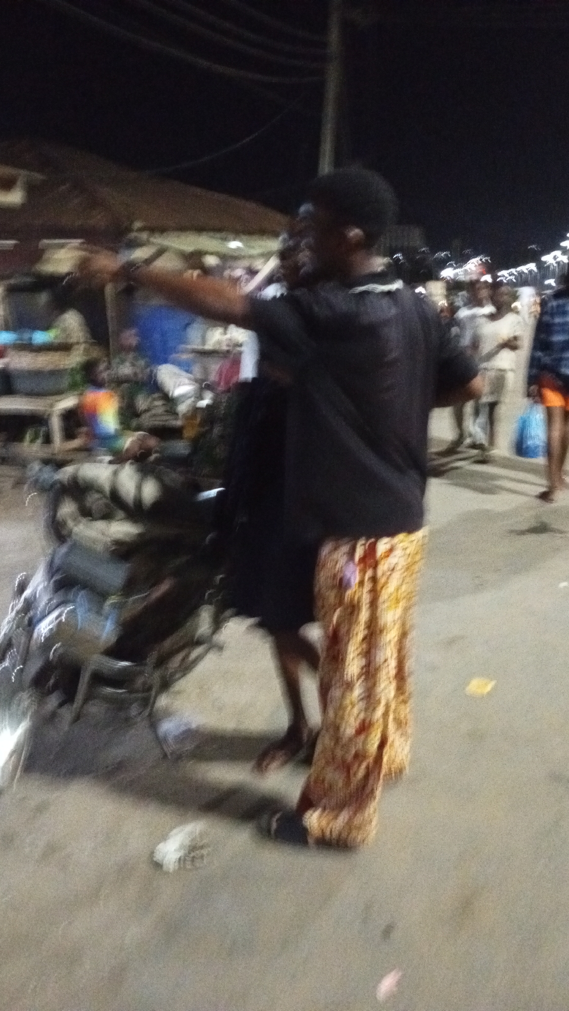 In Adankolo, Nigeria, at night, a person wearing a black t-shirt and patterned orange and brown pants points to the left. A motorcycle is parked next to the person. Other people are blurred in the background. Debris is on the ground. Buildings are visible in the background.