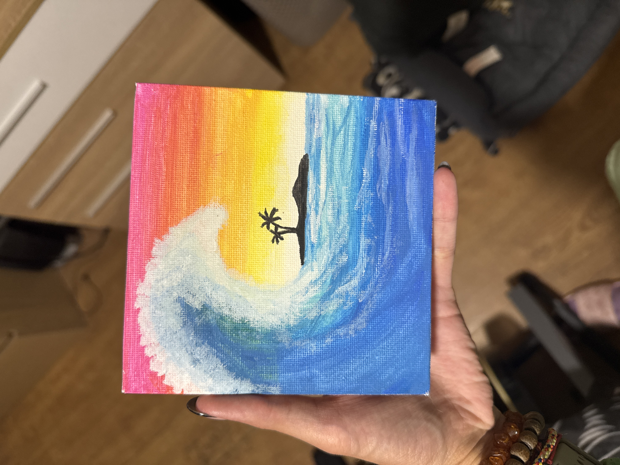 A square canvas painting is being held in someone's hand. The painting depicts a colorful scene, with pink, orange, and yellow blending on the left side, transitioning to blue on the right. A white wave with blue accents curves across the lower half of the painting. A black silhouette of a palm tree and rock formation is positioned in the center, against the yellow background. The background is out of focus and shows what appears to be a room with furniture. The specific location in Sofia, Bulgaria is not apparent.