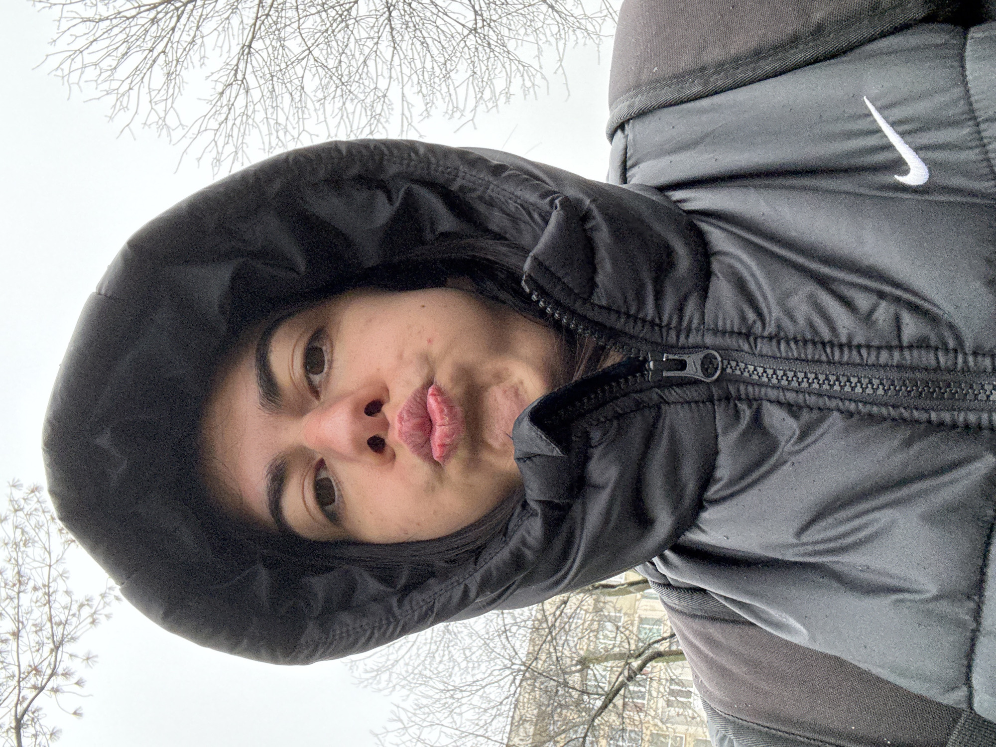 A close-up selfie-style photograph depicts an individual, likely an adult, from the chest upwards, outdoors in an urban area of Sofia, Bulgaria. The person is wearing a black hooded puffer jacket with the hood pulled up and zipped, partially obscuring the face. A white Nike "swoosh" logo is visible on the right chest area of the jacket, which exhibits small water droplets consistent with light precipitation or condensation. A black backpack strap is visible over the individual's left shoulder. The person has dark hair, brown eyes, and is making a "pout" expression with puckered lips. The background features a bright, overcast sky, indicative of dull weather, and numerous bare tree branches. A light-colored multi-story building with visible windows is partially seen through the branches on the left side of the frame, confirming an urban setting.