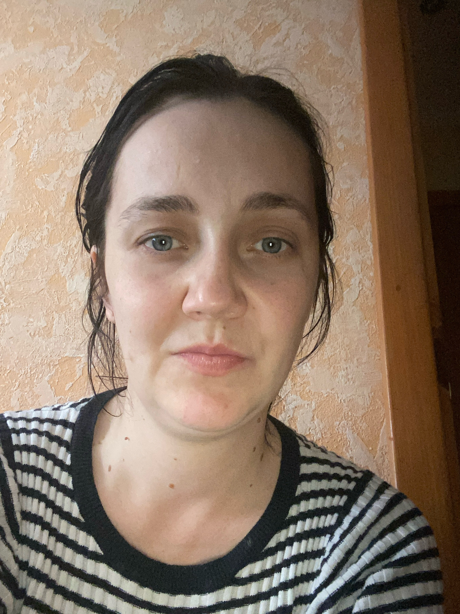 A medium shot shows a white woman with dark hair and blue eyes looking directly at the camera. She is wearing a black and white striped shirt. The background is a textured, peachy-colored wall. A brown wooden door frame is visible on the right side of the image. The lighting is natural and appears to be coming from the left.