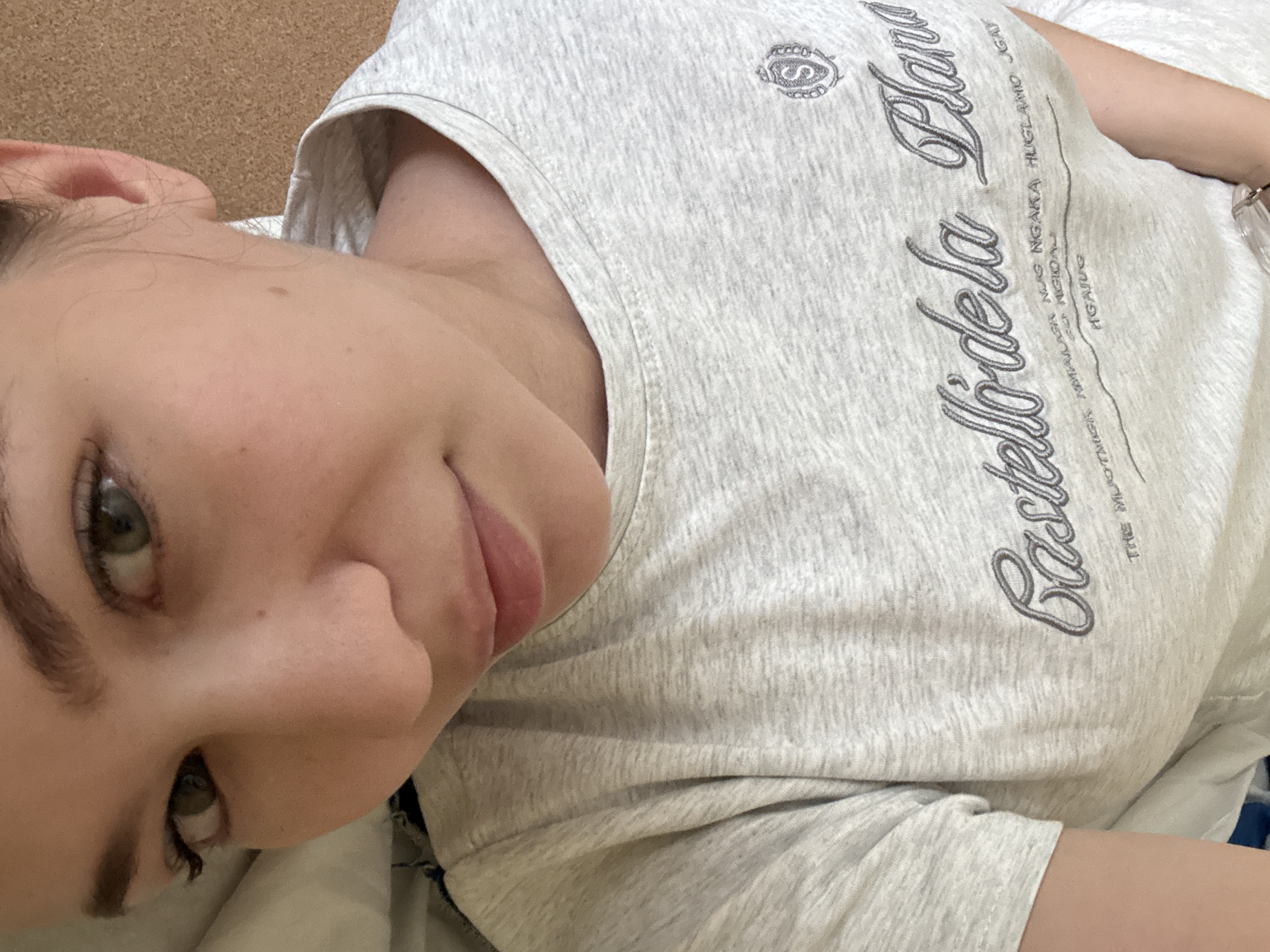 A single individual, appearing to be a young female, is depicted from a high-angle perspective, lying on a surface and looking directly towards the camera. The subject has fair skin, light-colored eyes (appearing green/hazel), defined eyebrows, and prominent dark eyelashes. Her lips are full and slightly parted. A small dark mole is visible on the right cheek. The subject wears a light grey, heathered long-sleeved shirt. The shirt features dark grey embroidered or printed text on the chest, prominently displaying "Castelladela Plana" in a script font. Above this, smaller, rotated text includes words such as "THE MODIFIER", "AMALAGA", "NGAGARA HUGLAM", "GAT", and "NGAJUQ". A stylized crest containing the letter "S" is visible on the upper left chest area of the shirt (viewer's right). The subject is in a supine or semi-supine position with her head slightly turned and eyes directed upwards. The image is rotated counter-clockwise. The setting appears to be indoors, with a light brown, textured surface (possibly cork) visible to the left of the subject's head, and white fabric (likely bedding) visible to the right and behind her head. The lighting is soft and ambient. The image provides no discernible visual information to determine its specific geographical location, including any connection to Chaikovsky, Russia.