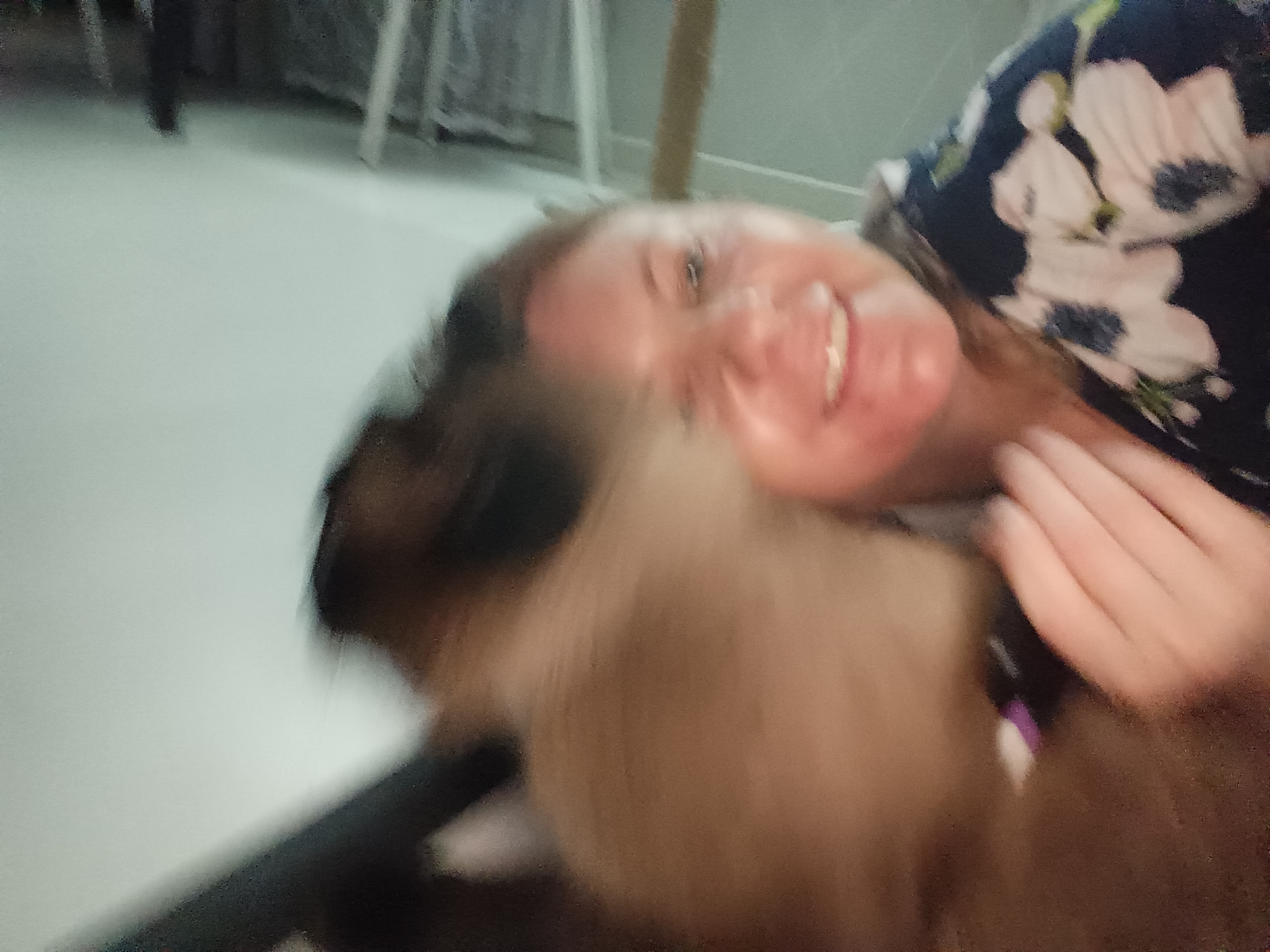 A young woman in a floral dress is lying on her side, looking up with a wide smile. A dog is in the foreground, its head close to the woman's face. The scene appears to be indoors, possibly a living room. The woman's hair is brown, and she has fair skin. The dog's fur is light brown and white. The background is blurred, showing a light-colored floor and some furniture. The image is taken from a low angle, suggesting the woman and dog are on the floor. There are no explicit indications of the image being taken in Sochi, Russia.