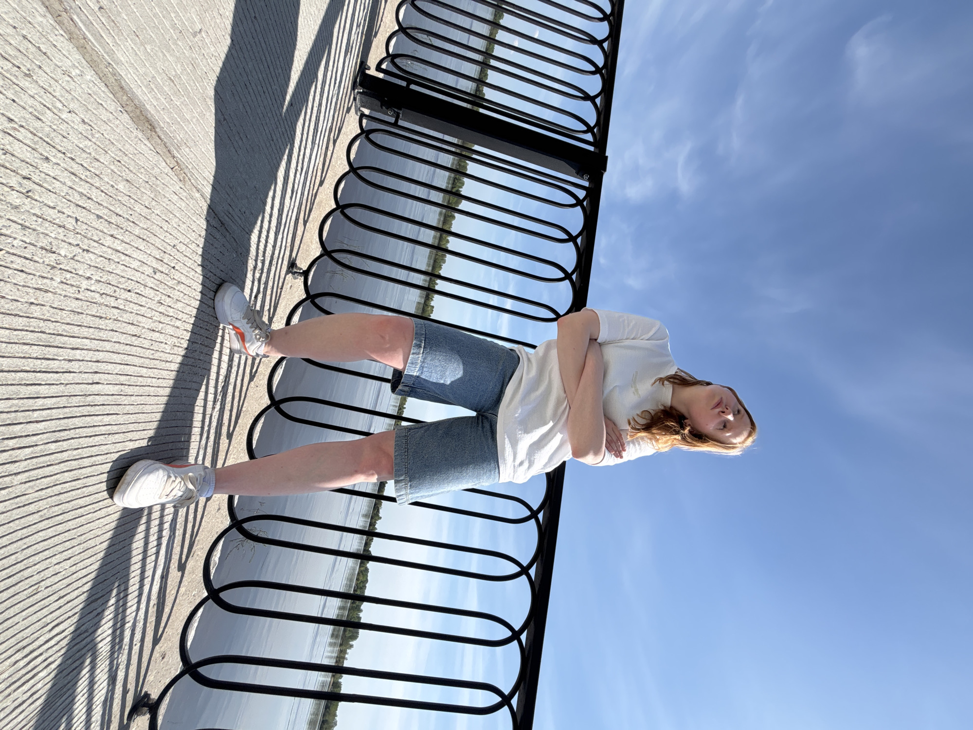 A full-length photograph features a person standing on a concrete walkway next to a black metal railing. The person has long brown hair and is wearing a white t-shirt, denim shorts, and white sneakers with orange accents. They have their arms crossed.
Beyond the railing is a body of water, which reflects the clear blue sky. The scene is brightly lit, indicating a sunny day.
The location is identifiable as the city of Perm, Russia, based on the characteristic design of the railing seen on the city's waterfront.