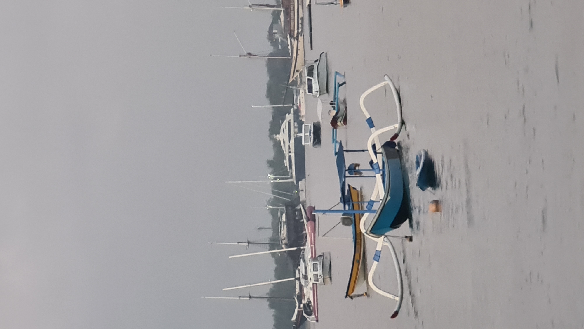 The image depicts a harbor filled with various boats under an overcast sky. In the foreground, a traditional Balinese outrigger boat, painted blue and white, is prominently featured. To its left, a smaller yellow and white boat is moored. Further back, several other vessels, including yachts and speedboats, are visible. In the far distance, a large, white luxury yacht with multiple decks stands out. The water appears calm with ripples, and the overall atmosphere suggests a cloudy or possibly rainy day. The scene is consistent with a coastal area or harbor in Denpasar, Indonesia, likely a place where boats are docked or anchored. There are no people clearly visible in the image.