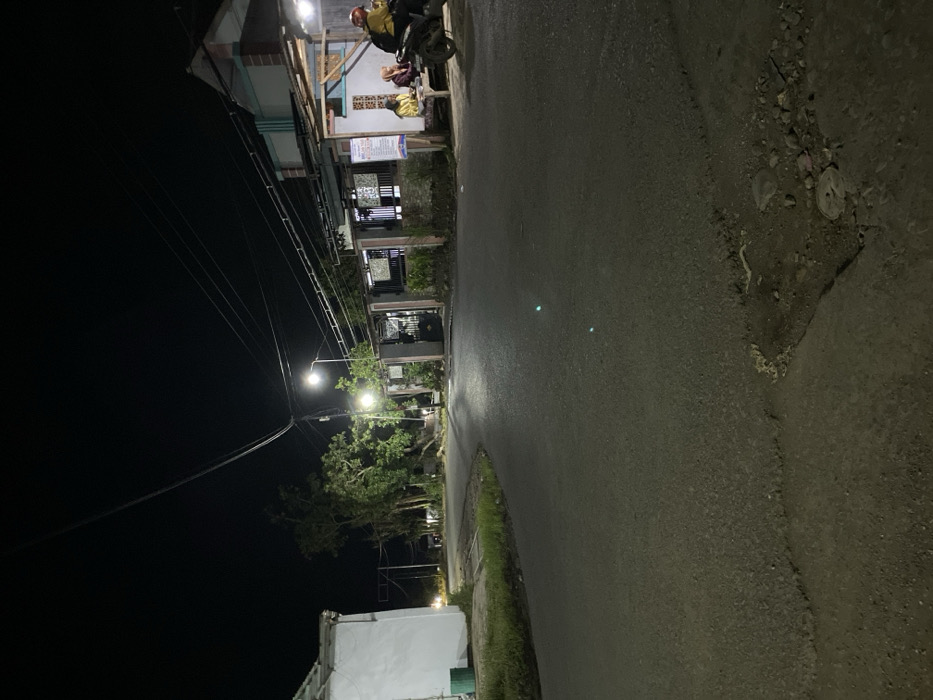 The image depicts a nighttime street scene in Gading Kulon, Indonesia. An asphalt road, showing signs of wear and minor potholes in the foreground, curves towards the middle-ground, illuminated by multiple overhead streetlights. On the left, a portion of a white building with a green trash can is visible, adjacent to a patch of grass. Further down this side, a pedestrian is faintly visible walking near a cluster of trees. On the right side of the road, a row of residential or commercial buildings is present. One building features a brightly lit, open-air area with a shelter. Here, three individuals are seated at a table, two of whom are facing each other, possibly interacting or eating. Adjacent to them, a person in a yellow jacket and red helmet is positioned on a parked dark-colored scooter or motorcycle. Utility poles and a dense network of overhead power lines span across the upper portion of the frame, connecting to the structures. The overall scene is lit by artificial light sources, contrasting with the dark night sky.