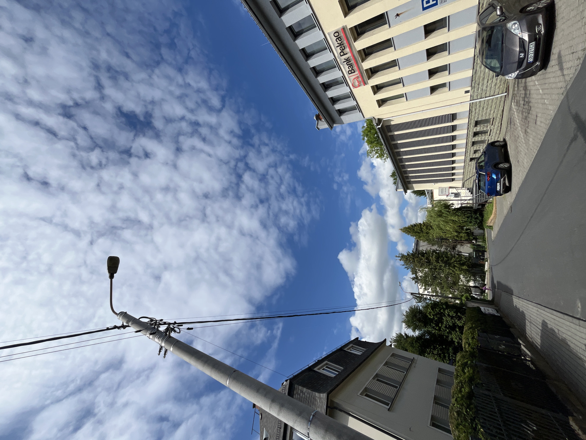 The image, when viewed upright, presents a sunlit urban street scene under a bright blue sky with scattered white cumulus clouds.
In the foreground on the left, a tall, grey concrete lamppost with attached power lines extends diagonally upwards and across the frame. To its right, a multi-story building with a dark, sloped roof and light-colored walls is partially visible.
On the opposite side of the street, a large, prominent building with a light yellow facade and grey window frames features a striking red and white sign that clearly reads "Bank Pekao". A blue "P" sign, indicating parking, is also visible on this building. Below the main facade, a section of the building exhibits a pattern of horizontal grey louvers.
Two cars are parked on the street next to the Bank Pekao building. The first car, a dark grey/brown SUV (appears to be a Honda CR-V), has a license plate "SB 22TGY". Behind it, a blue SUV is parked, with the license plate "UB384HM". The street is paved, and a sidewalk runs along the right side.
Green trees and other foliage are visible along the street and in the background, adding natural elements to the urban setting. The bright sunlight casts distinct shadows, suggesting it is daytime, likely mid-day. No people or specific activities are visible in the scene. The "Bank Pekao" signage and the license plate formats indicate a location in Poland.