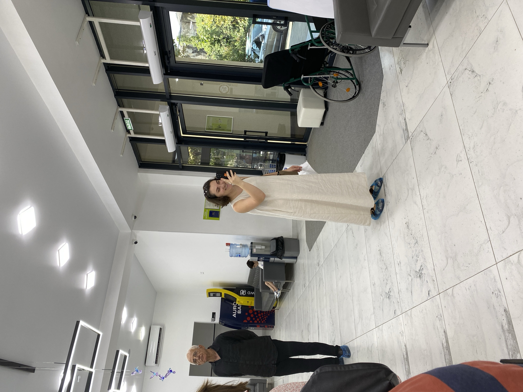 In a bright, modern interior space, likely a clinic or medical facility in Sokhumi, Abkhazia, a woman in a long, cream-colored dress is holding up her phone, seemingly taking a picture or video of herself and the surroundings. She is standing in the foreground, facing right, with a slight smile on her face. To her left, a man in a black shirt and pants stands facing forward, with blue shoe covers on his feet. Behind him, a yellow ATM-like machine with red circular accents is visible, along with a seating area where a young boy sits on a grey couch. Further in the background, an empty wheelchair is positioned near a doorway, with a building visible through the glass. The floor is tiled with a light marble pattern.