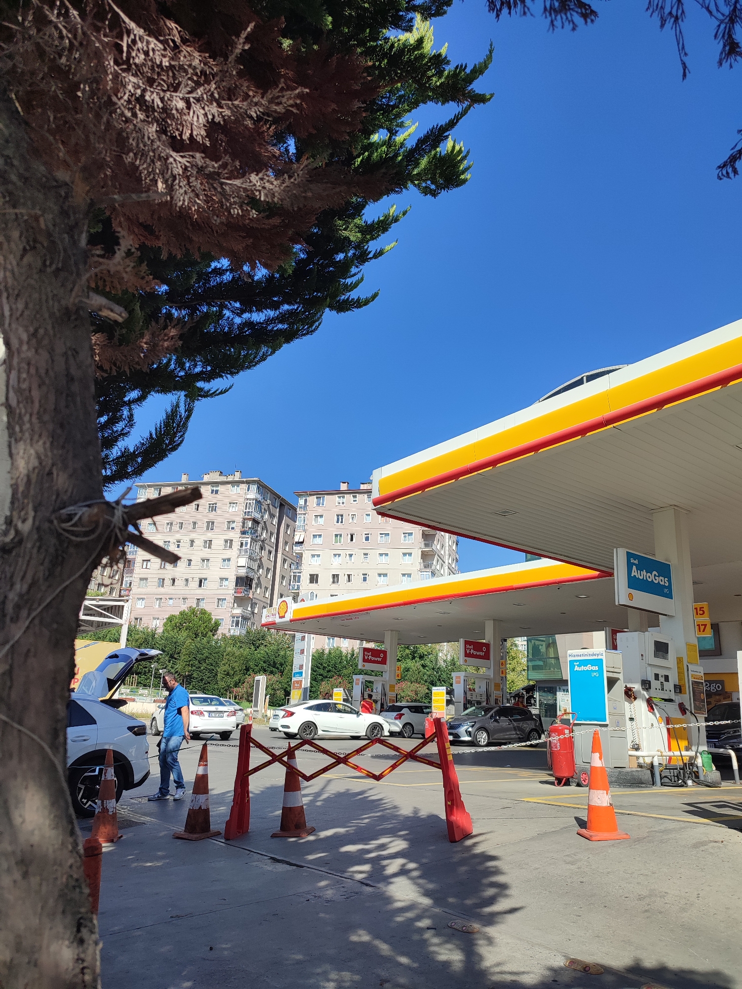 The image depicts a Shell gas station on a sunny day in Üsküdar, Turkey. In the foreground, a large tree trunk partially obscures the view on the left. The gas station canopy, with its distinctive yellow and red Shell branding, is prominent. Several cars are present at the pumps, including a white vehicle with its trunk open and a man in a blue shirt and jeans standing next to it. Orange traffic cones and a red foldable barrier are positioned on the ground, likely to manage traffic flow or indicate a closed area. In the background, multi-story apartment buildings rise against a clear blue sky. The scene conveys a typical urban setting with daily activities at a service station.