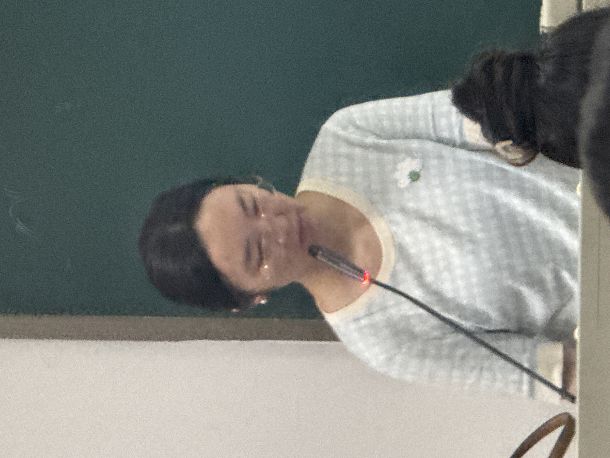 A woman with dark hair is speaking into a microphone in a room with a green wall and a white surface. She is wearing a light blue sweater with a white floral pin. The microphone is black with a red light on the top. Yuzhong District, China cannot be confirmed from the image.