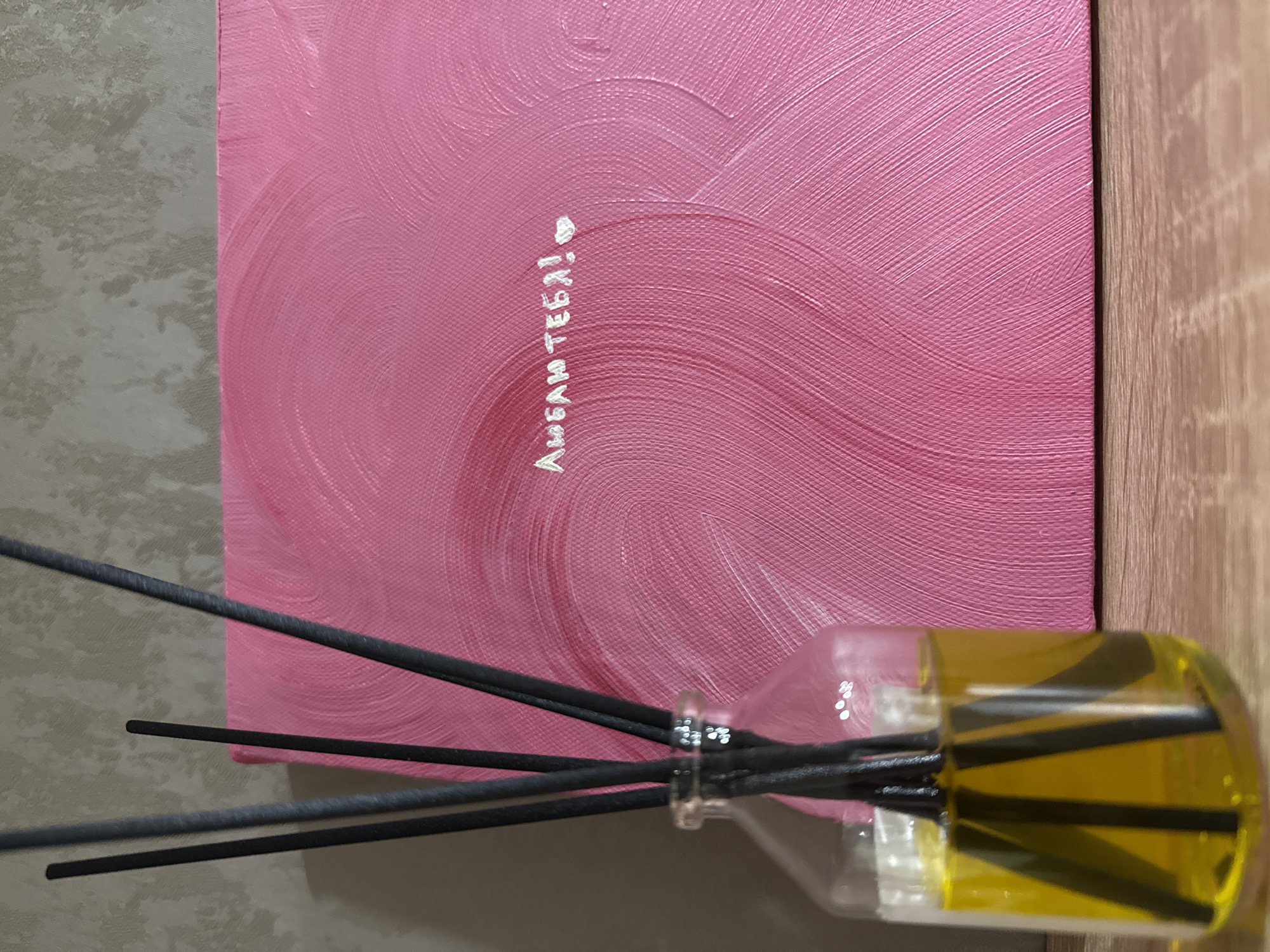 The image shows a still life composed of a small canvas and a fragrance diffuser. The canvas, positioned to the left, is painted in shades of pink with visible brushstrokes creating a swirling pattern. White lettering and a heart symbol are painted on the canvas. To the right, partially overlapping the canvas, is a clear glass bottle containing a yellow liquid. Several black reed sticks are inserted into the bottle, partially submerged in the liquid and extending upwards. The backdrop includes a light grey surface in the lower left and a light brown, wood-grain surface on the right. The origin of the image is the city of Chaikovsky, Russia.