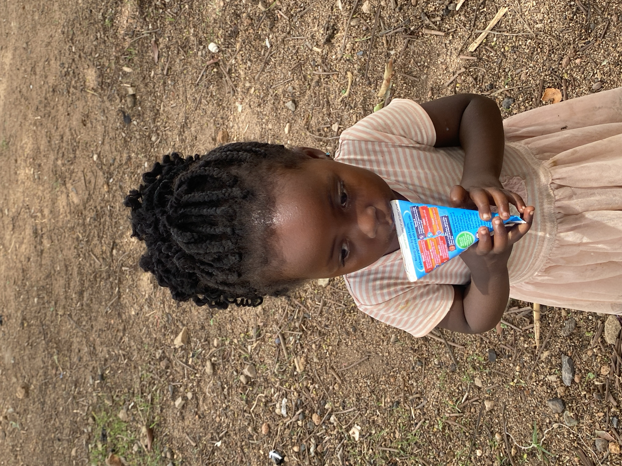 The image captures a child with braided hair, dressed in a light pink striped top and a tulle skirt. She is standing on ground scattered with small stones and twigs, likely in the city of Kuru Dungus, Nigeria. The child is holding a blue and white carton drink labeled FreshYo, suggesting she may be drinking or about to drink it. The image is taken from a high angle, offering a clear view of the child and the ground around her.