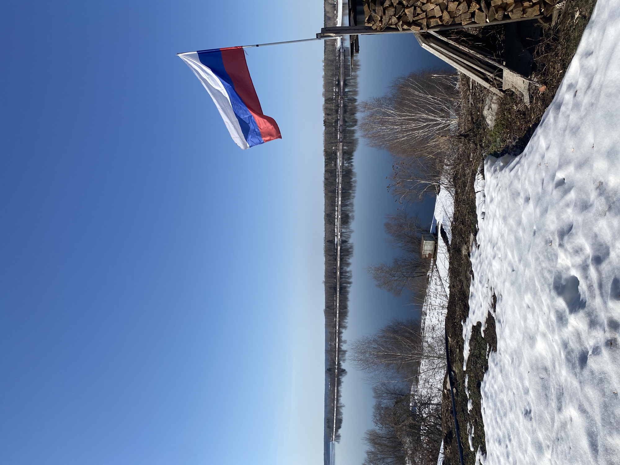 The image shows the Russian flag waving in the wind against a clear blue sky in Olkhovka, Russia. Beneath the flag, a body of water stretches into the distance, its surface reflecting the sky. The shoreline is visible on the right side of the image, covered in snow and dotted with trees. A small structure is partially visible near the top right, with a stack of wood beside it.